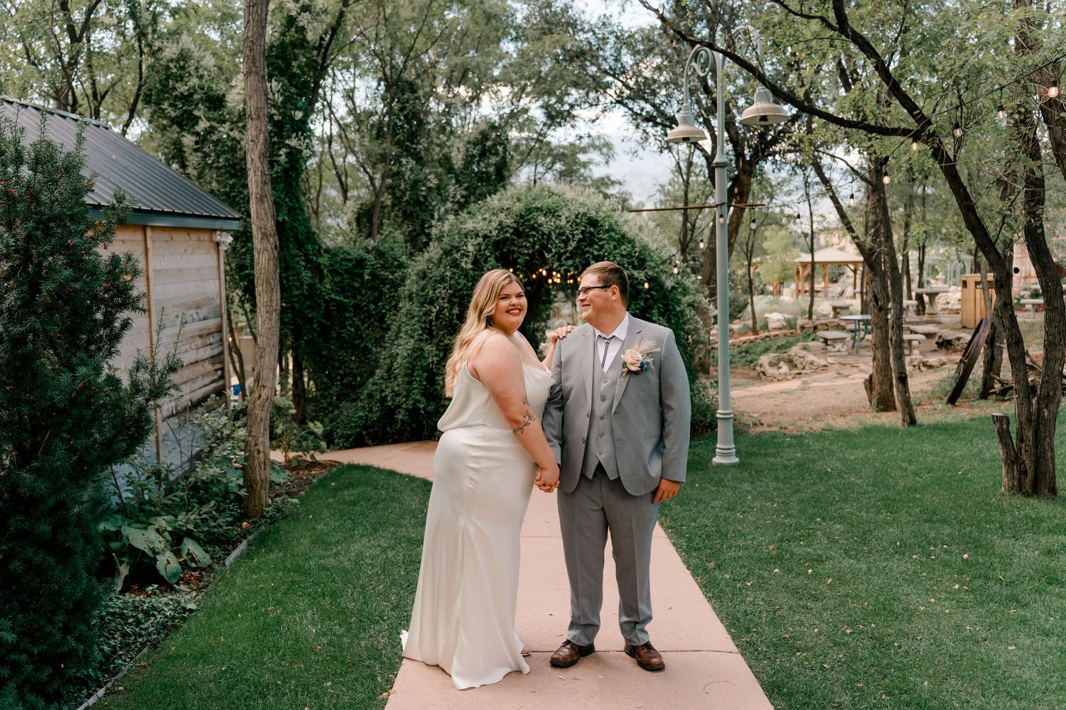 A couple in wedding attire stands together on a garden path surrounded by trees and greenery.