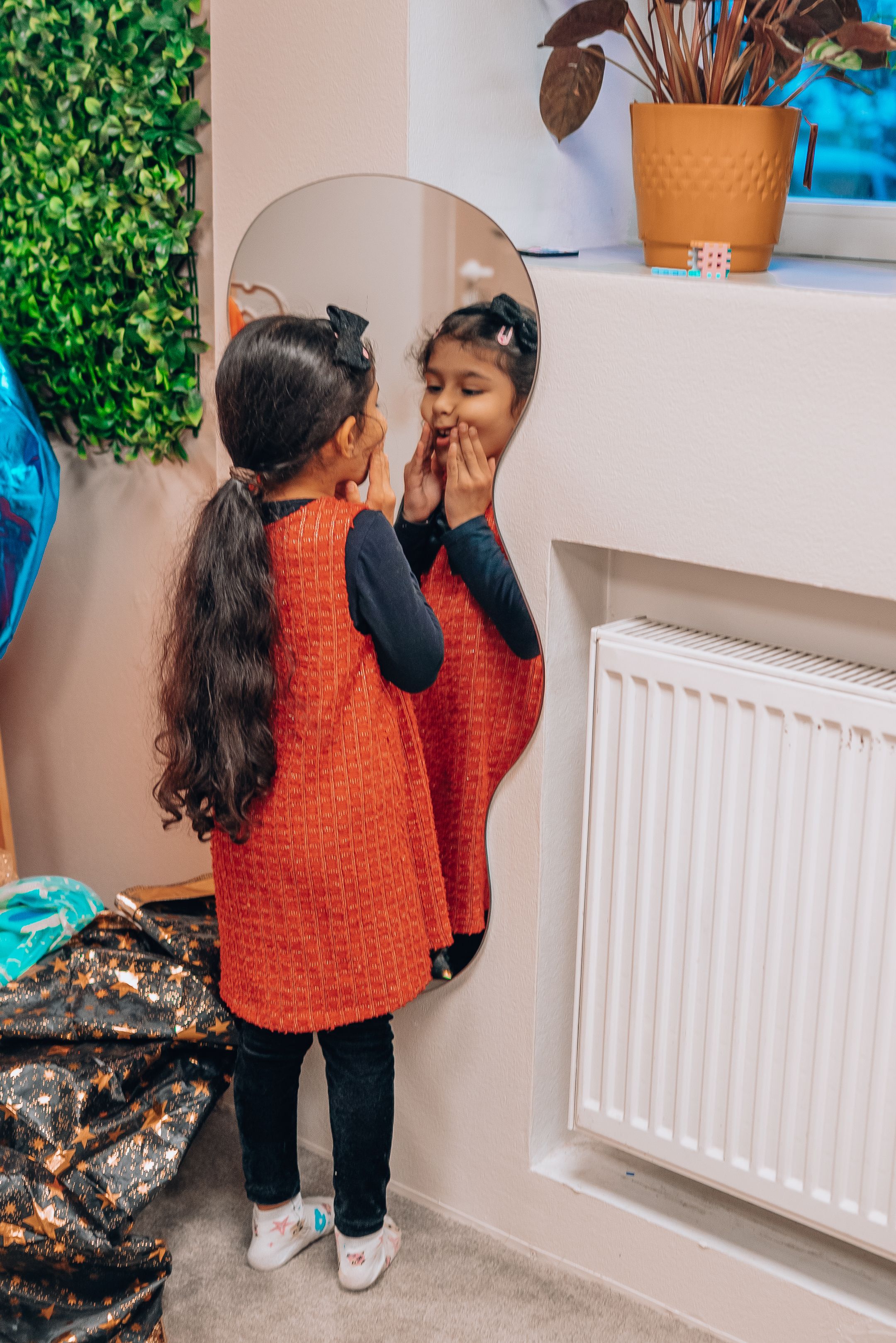 A person wearing an orange knit sweater and black pants reaches up to look at items on a white shelf near a radiator.