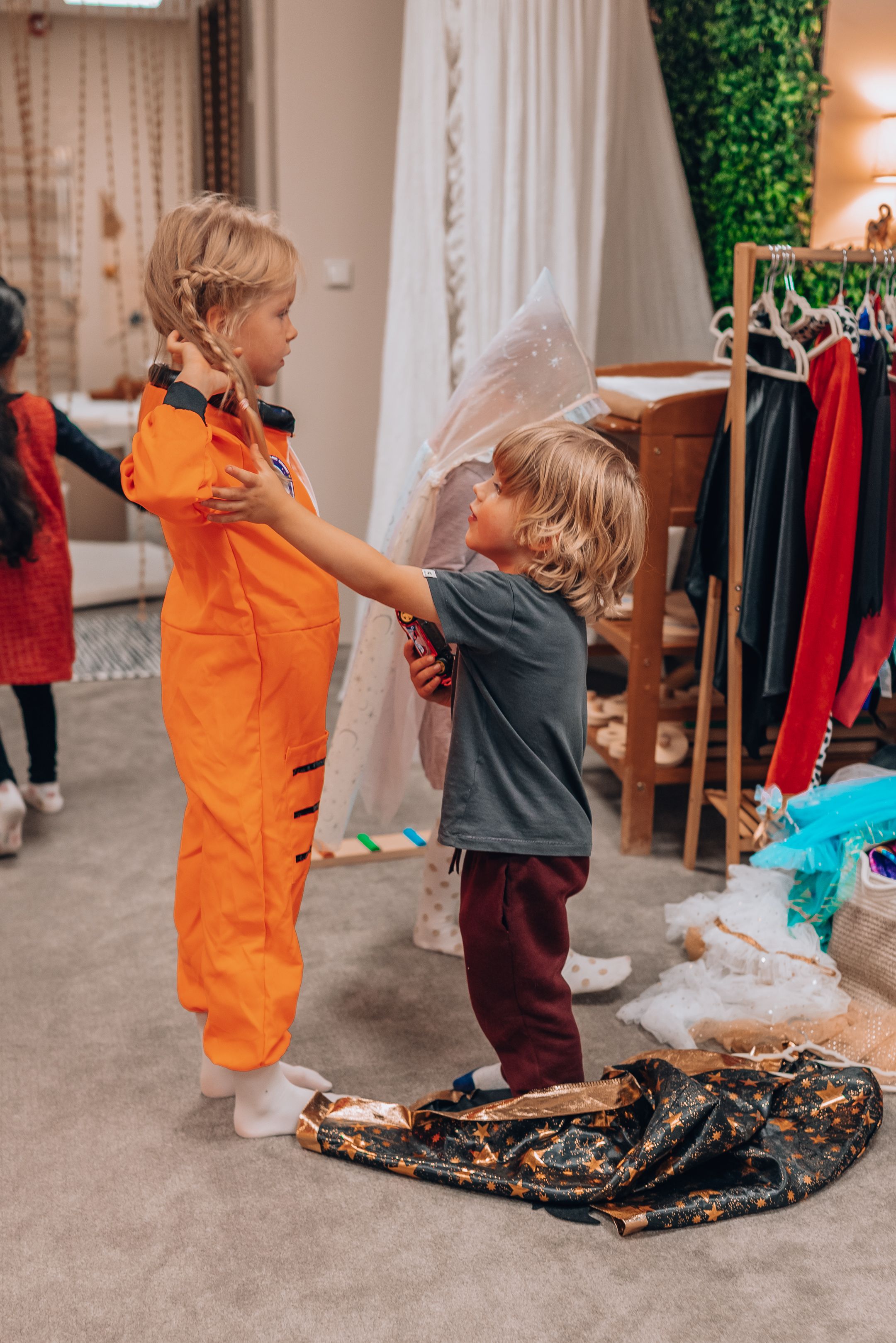Two children play with a sequined black mermaid tail blanket in a bedroom while surrounded by clothing.