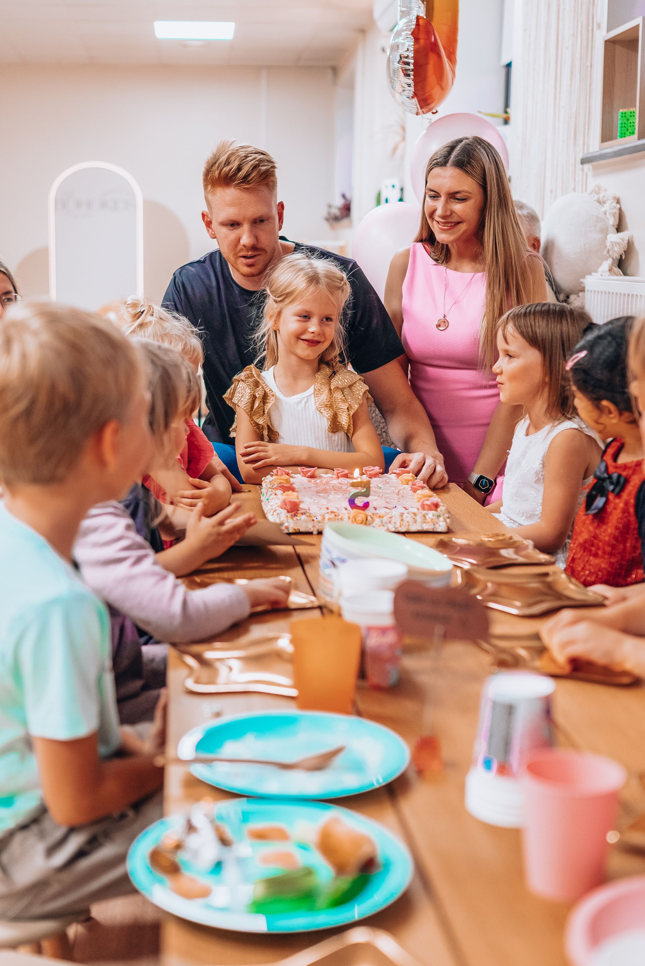 Children enjoying a birthday party celebration at a table with colorful plates and drinks while adults supervise the festivities.