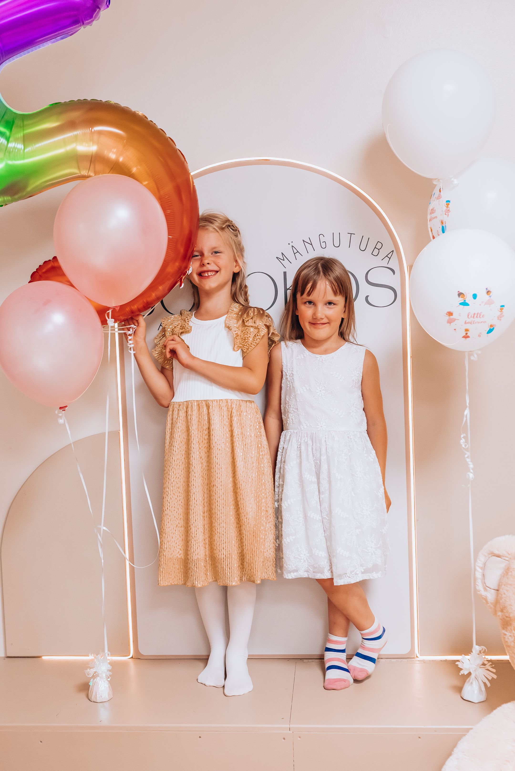 Children in white and gold dresses pose playfully with colorful balloons against a neutral wall with arched details.