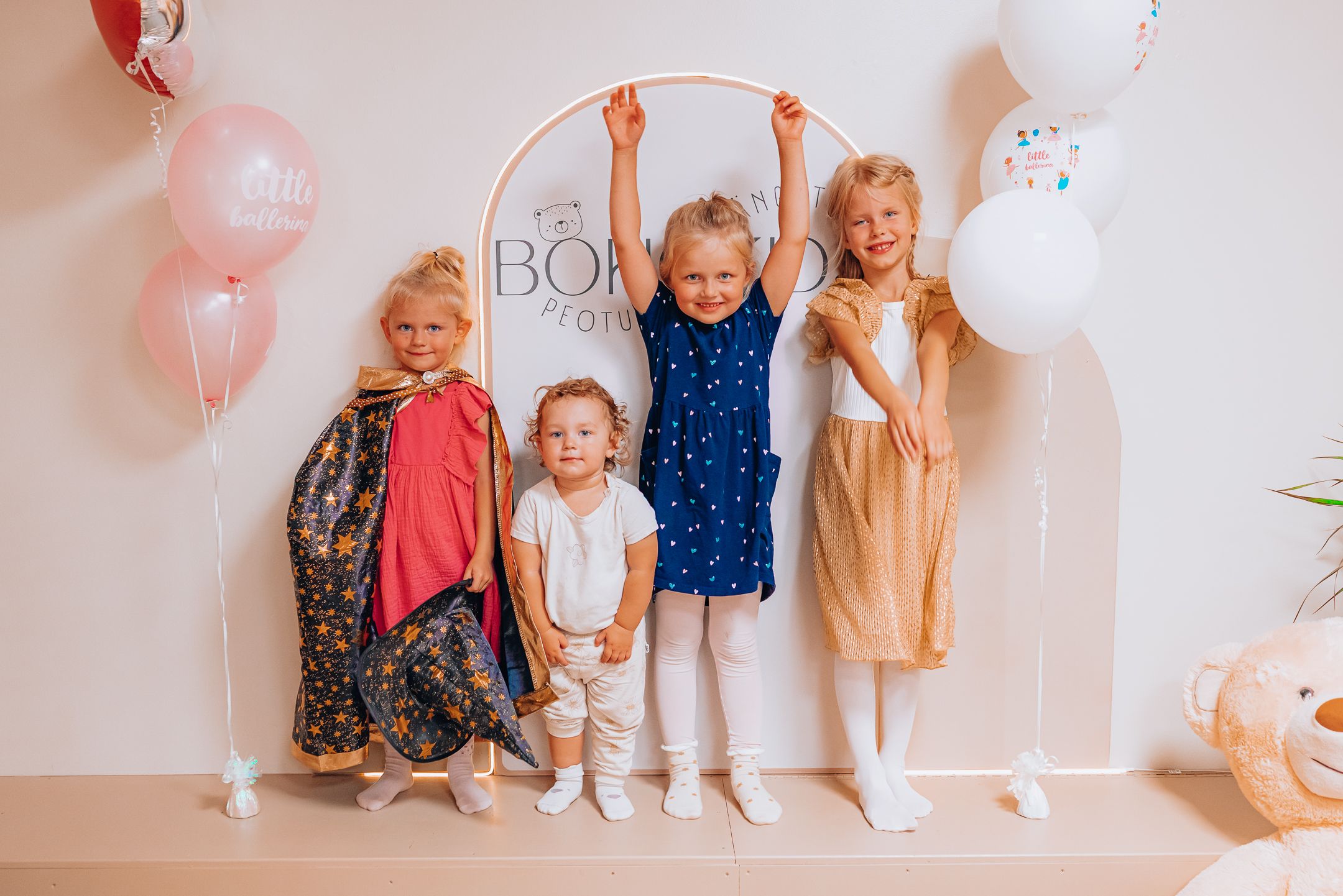 Children stand against a white wall with pink balloons creating a festive party atmosphere.