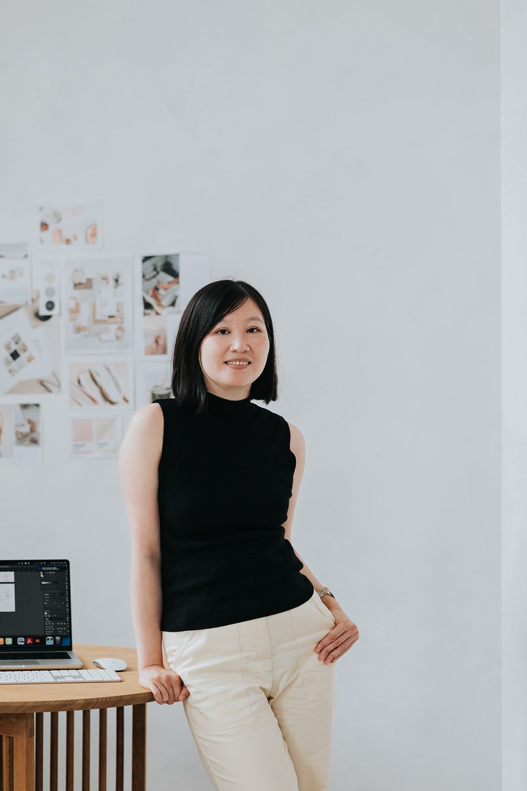 A person in a black sleeveless top and beige pants poses against a white wall with photos displayed in the background.
