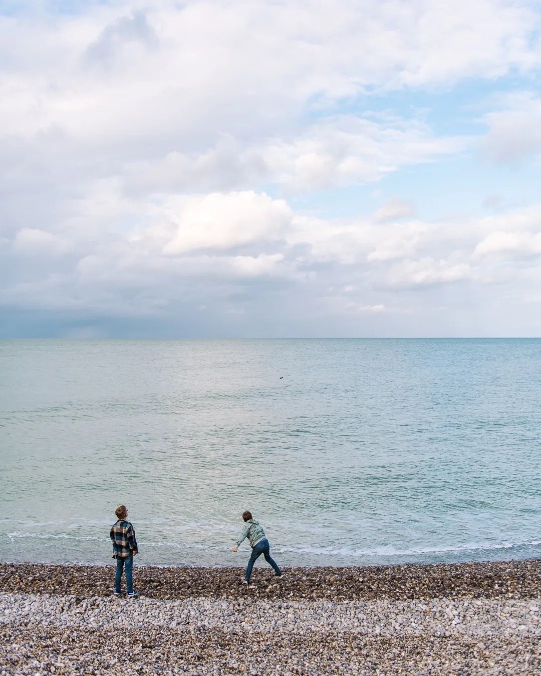 Two children walking along a pebble beach beside the calm sea on a cloudy day.