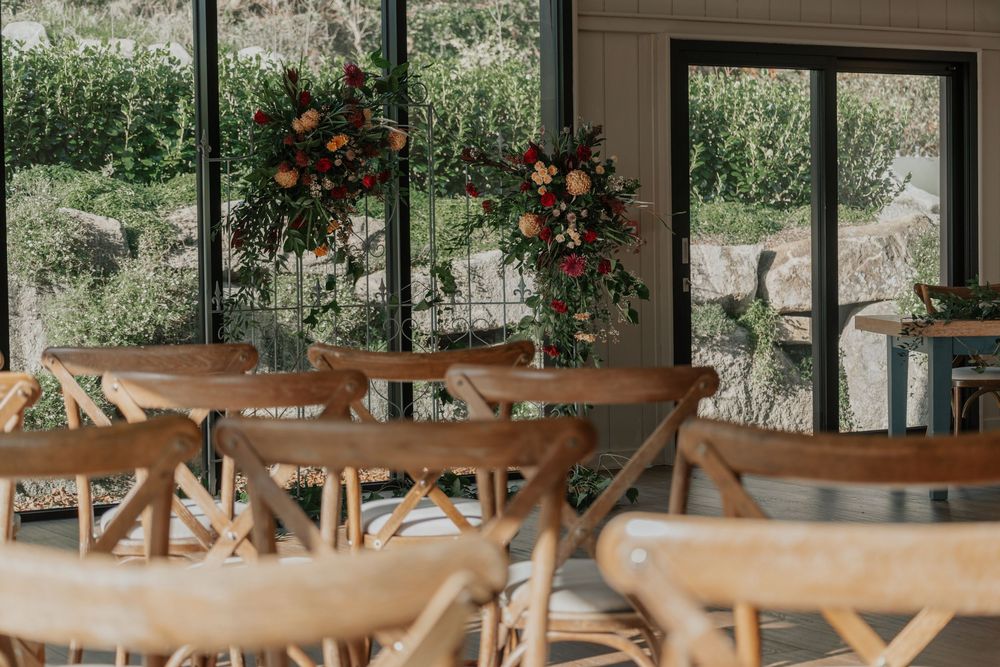 Rustic wedding ceremony setup with wooden cross-back chairs and vibrant floral arrangements by windows.