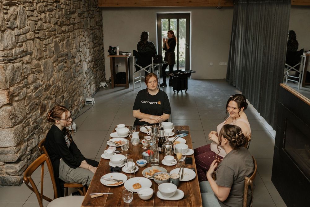 Wedding party guests seated around a rustic wooden table in a stone-walled venue during bridal prep.