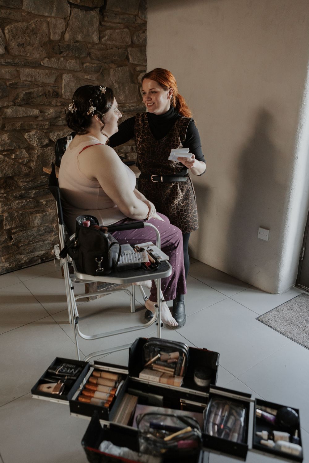 Makeup artist consulting with bride during wedding preparation in a rustic stone-walled bridal suite.