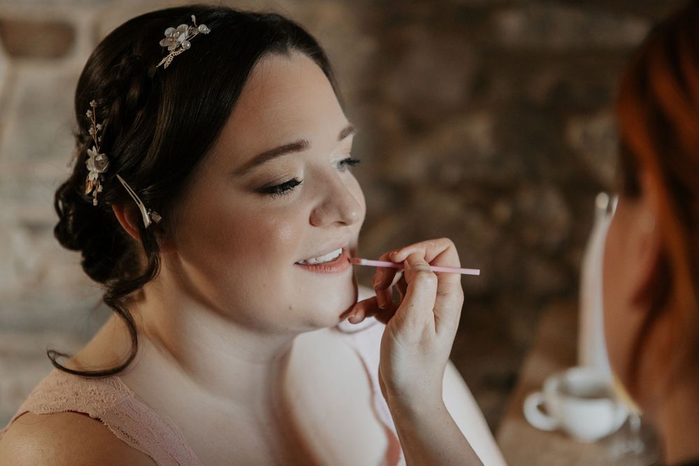 Professional makeup artist applying lipstick to a smiling bride with elegant updo hairstyle.
