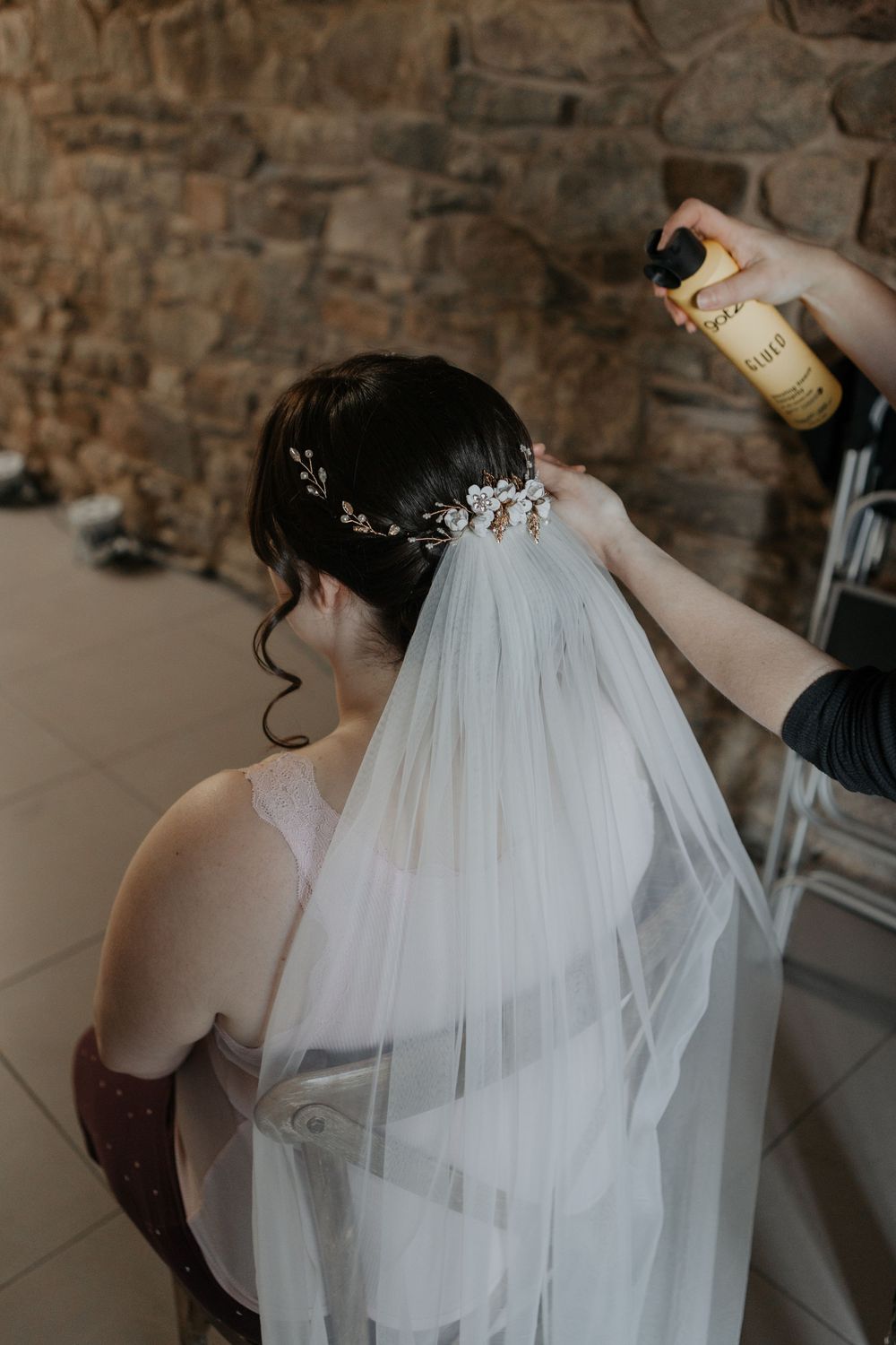 Hairstylist spraying and arranging a floral hair comb on bride's veil and updo before wedding.