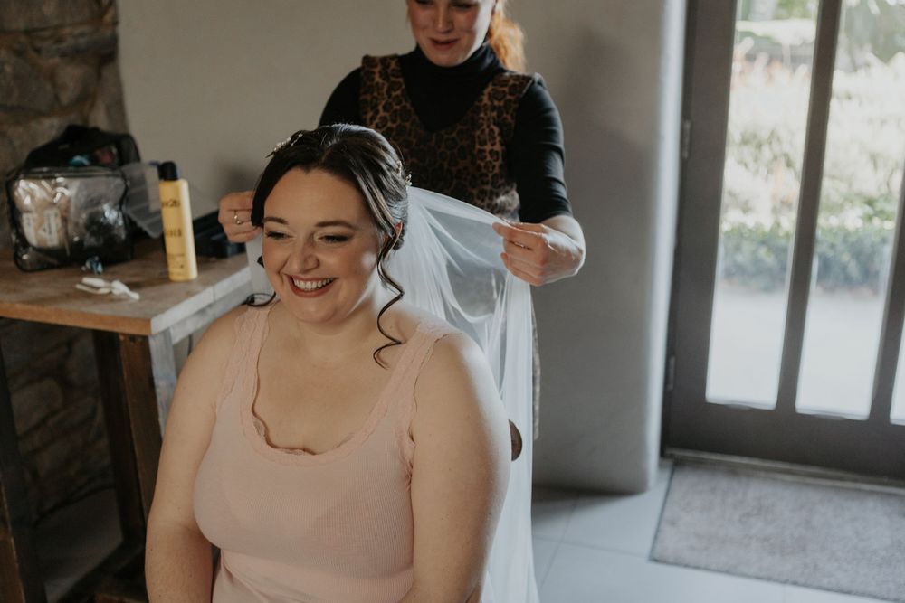 Smiling bride having her wedding veil fitted by hairstylist in bright bridal preparation room.