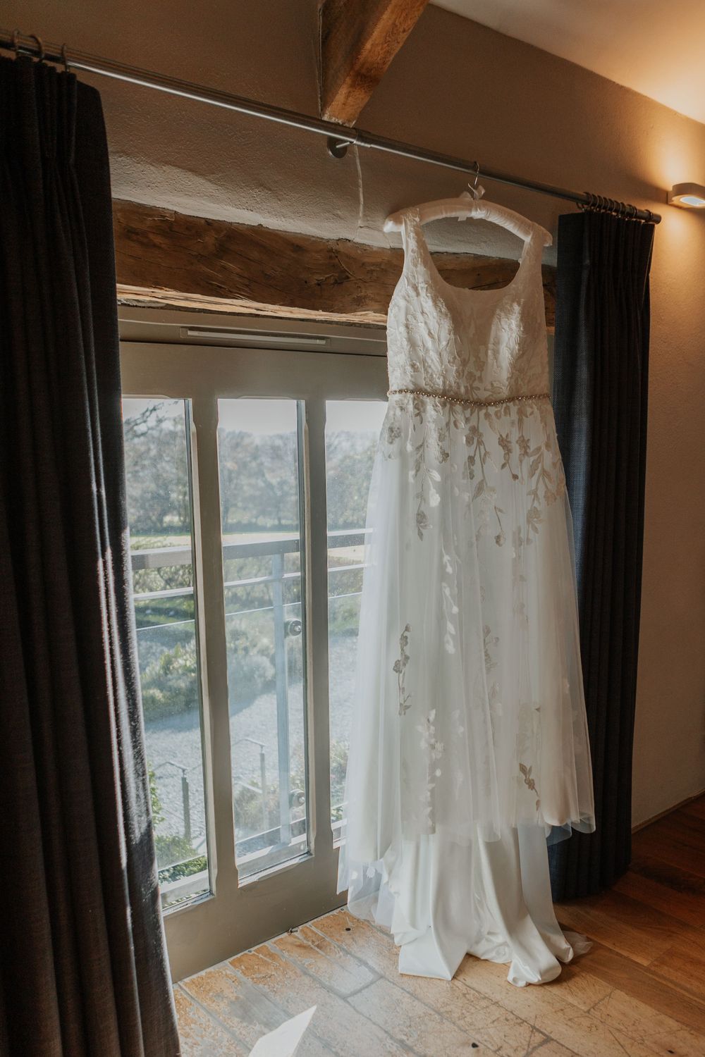 Elegant white lace wedding dress hanging by a window with natural light in a rustic barn venue.