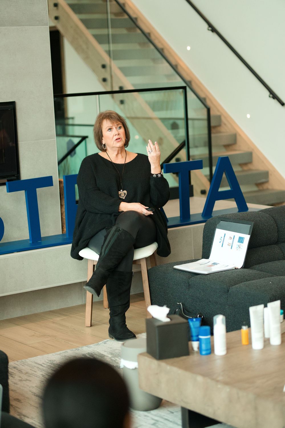 Speaker in black attire gestures while seated on a chair during a presentation in modern office space with stairs.