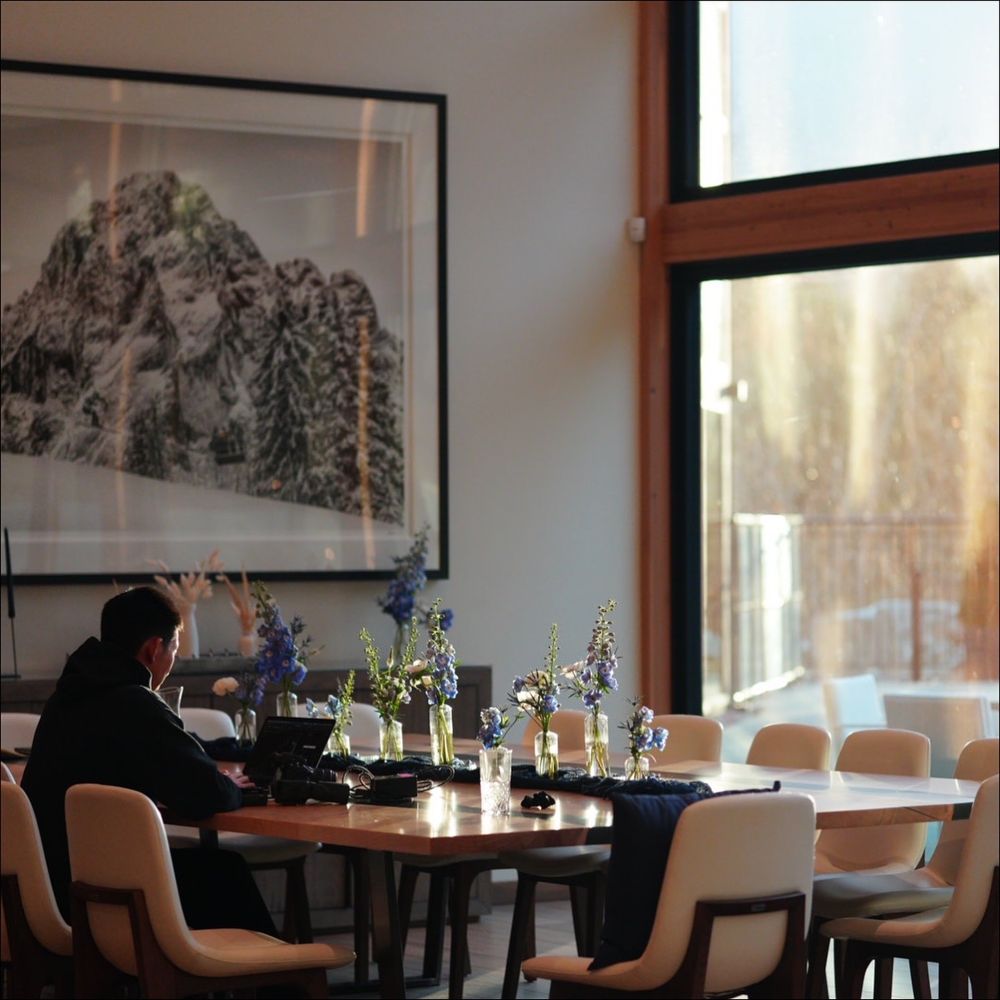 Modern restaurant interior with cream-colored chairs and wooden tables set against large windows with mountain views.