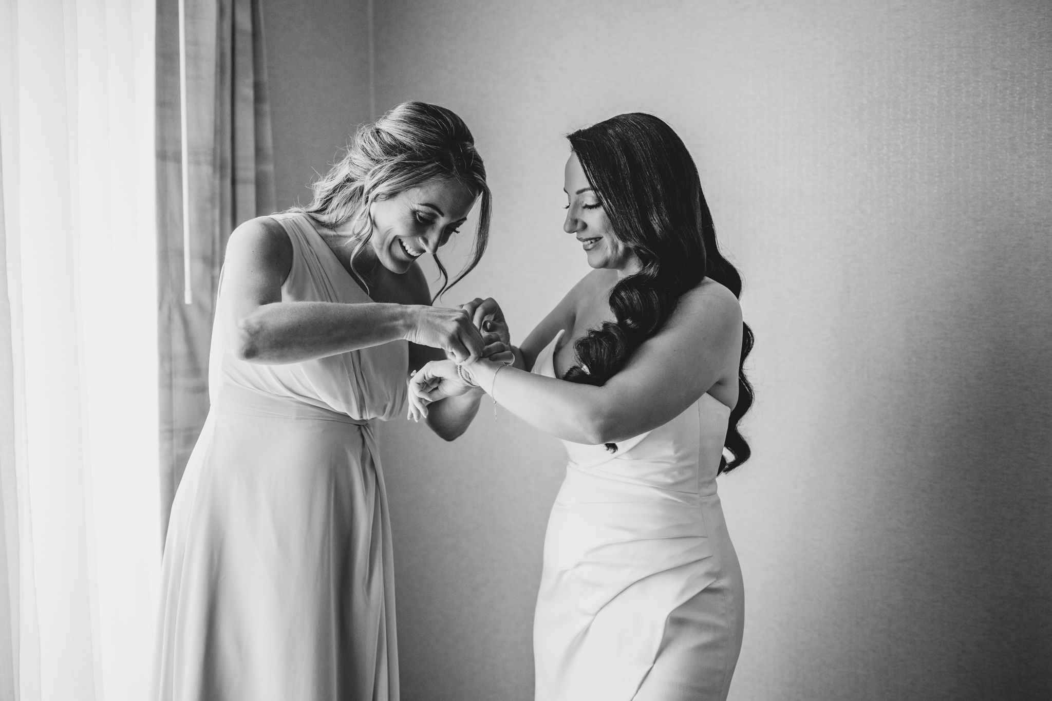 Two women in white dresses share a joyful moment while getting ready for a wedding.