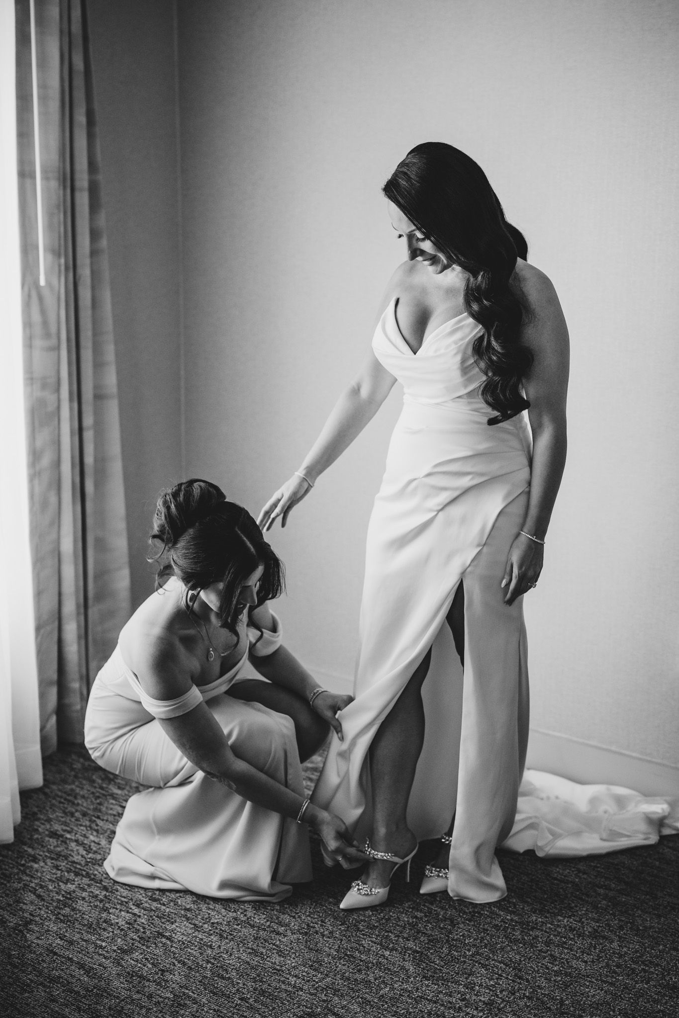 A bridesmaid helps adjust the bride's wedding dress and shoes near a window.