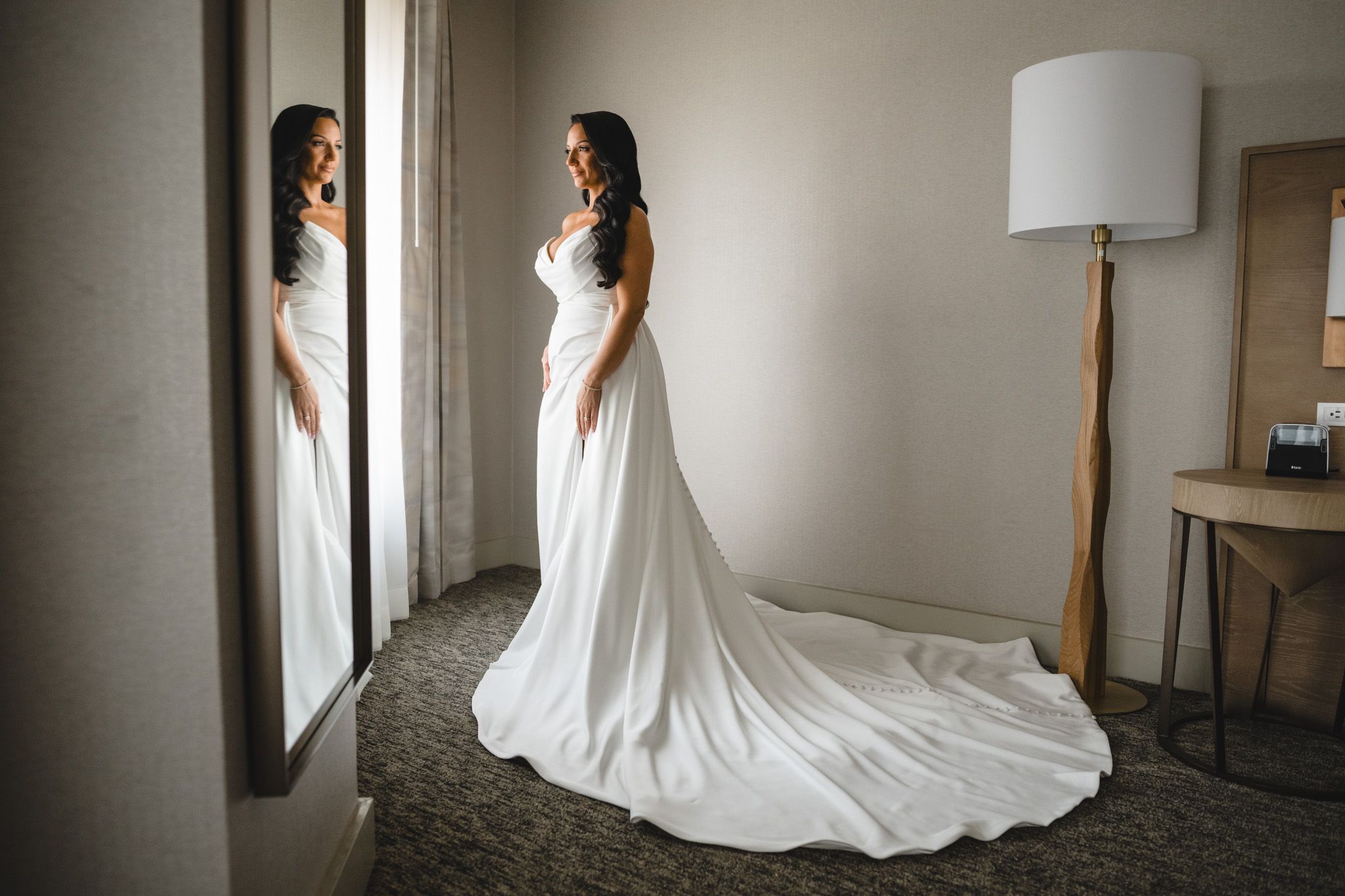 Elegant bride in a flowing white gown with long train stands by window in natural light.