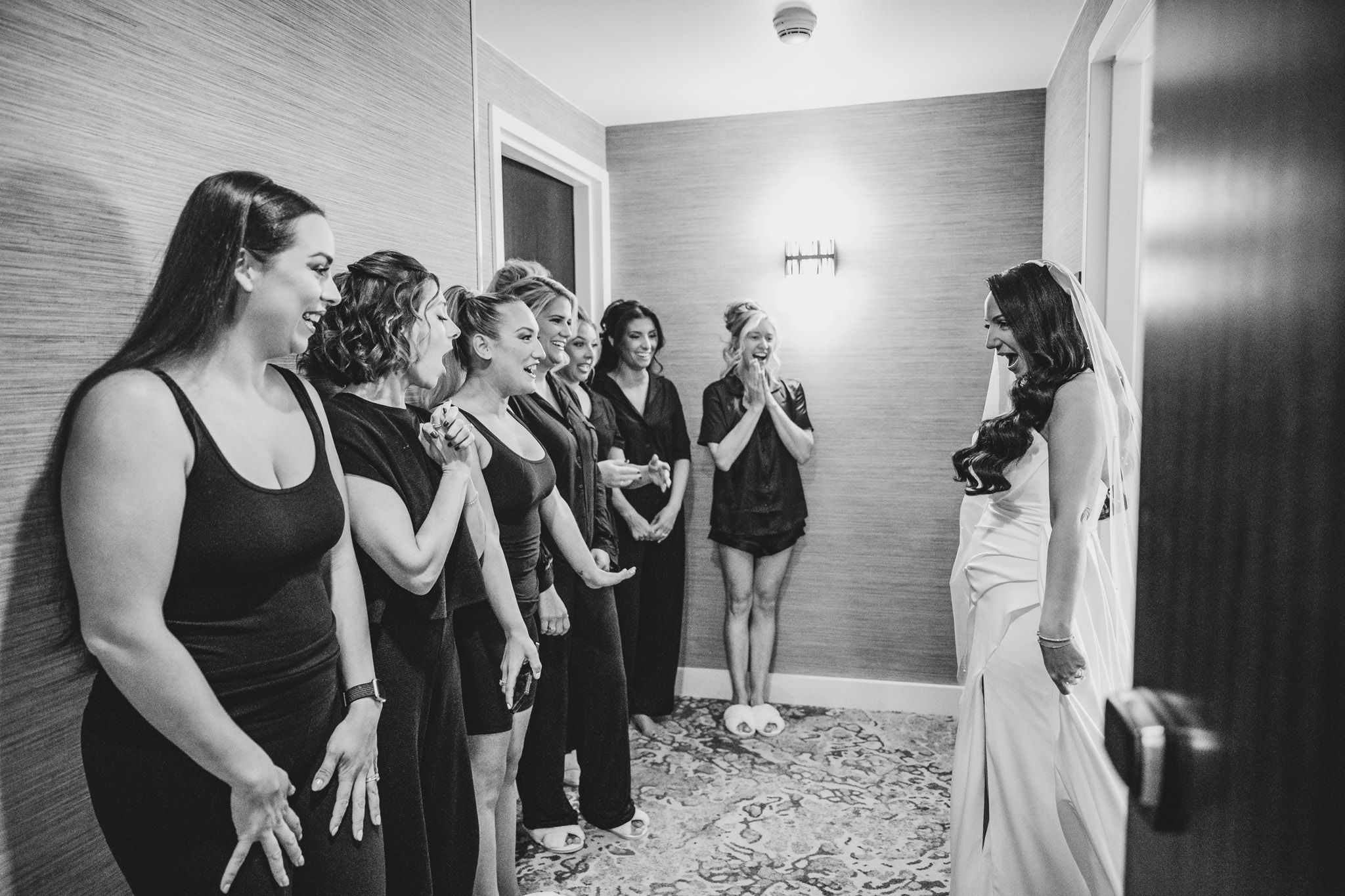 Wedding party in black dresses reacts with excitement upon seeing the bride.