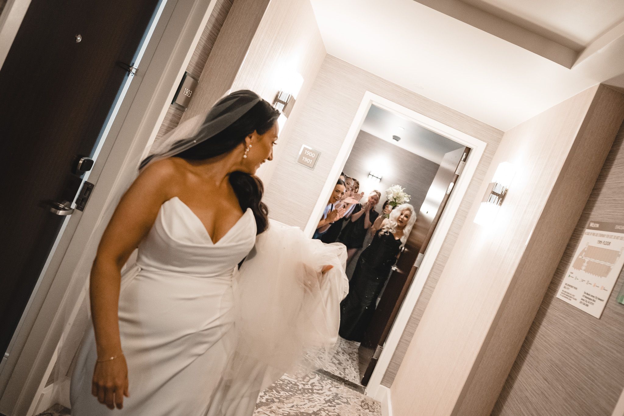 Bride in white gown reveals her dress to wedding party waiting in hallway.