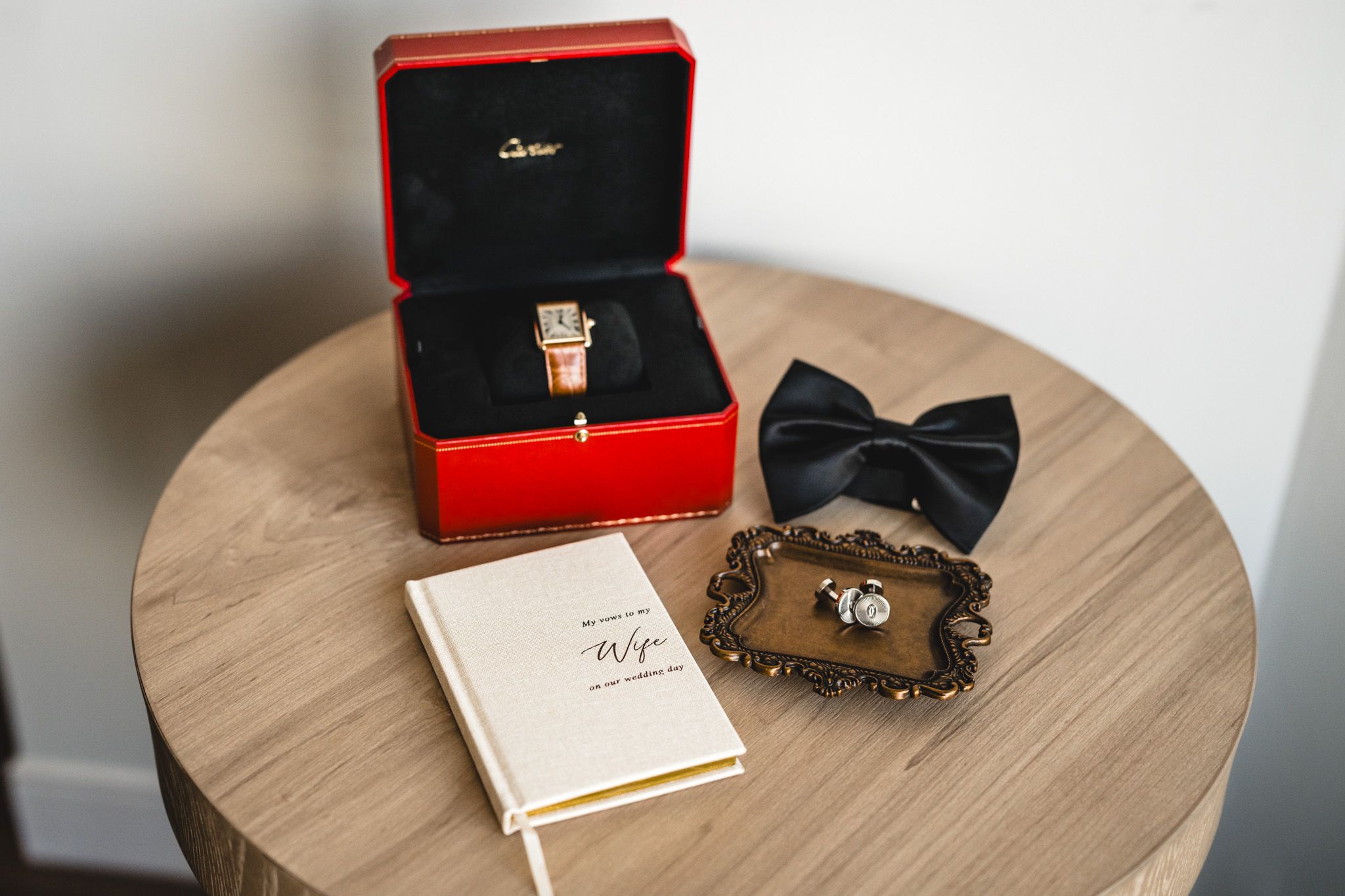 Wedding accessories including red ring box, black bowtie and vows book on wooden table.