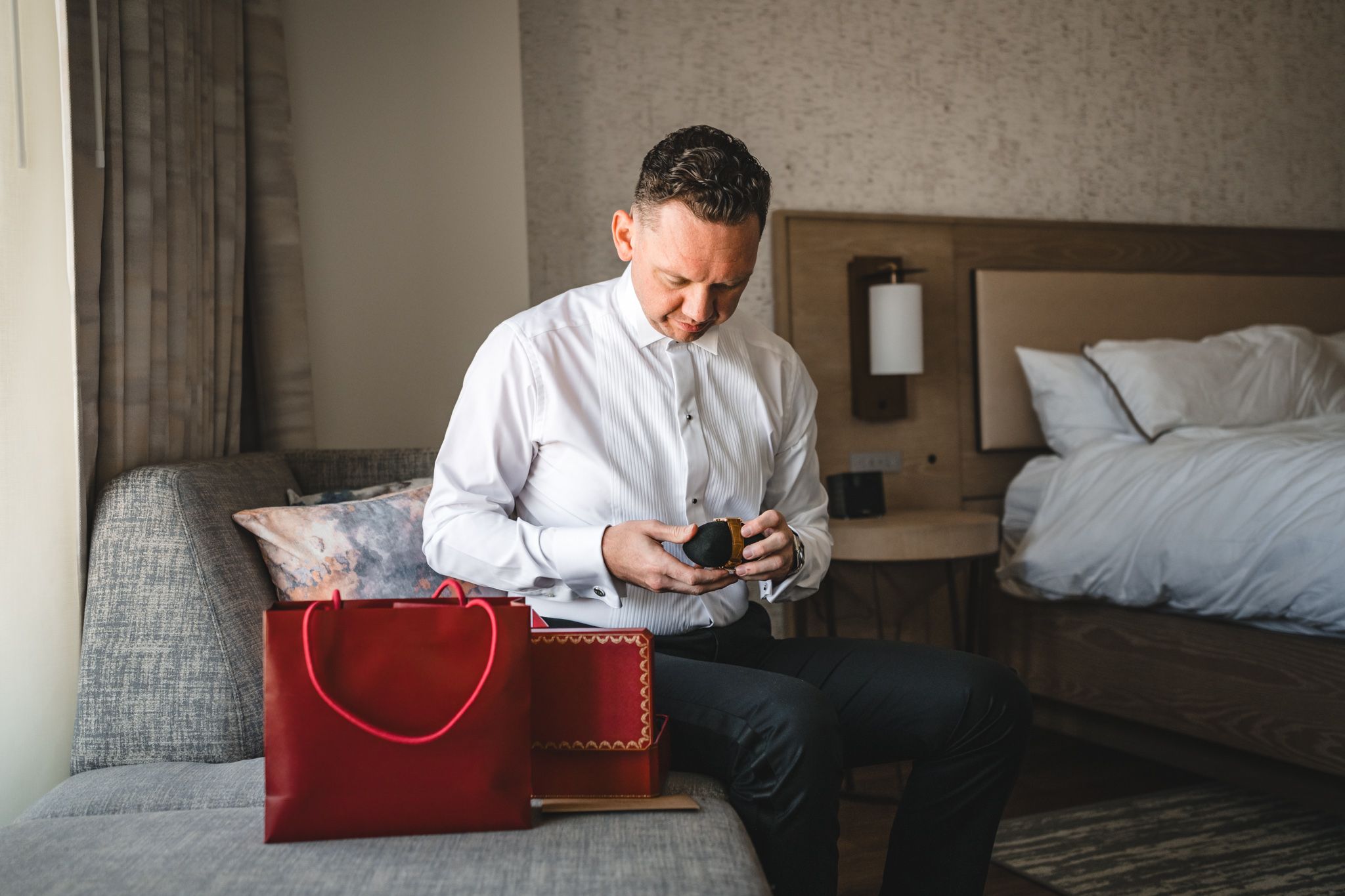 A person in a white dress shirt sits on a bed while looking at something in their hands.