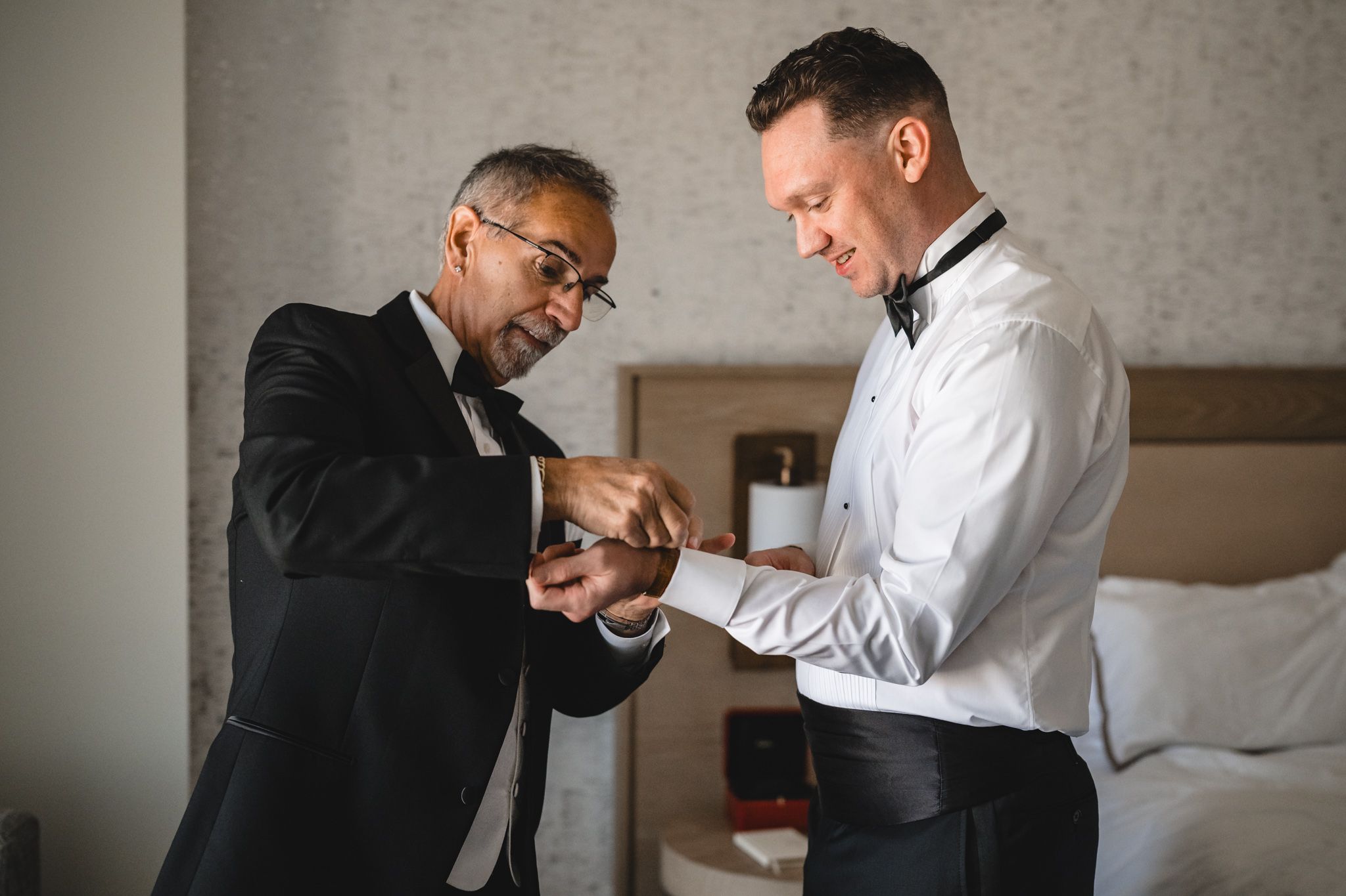 Two people in formal wear assist each other with adjusting their attire.
