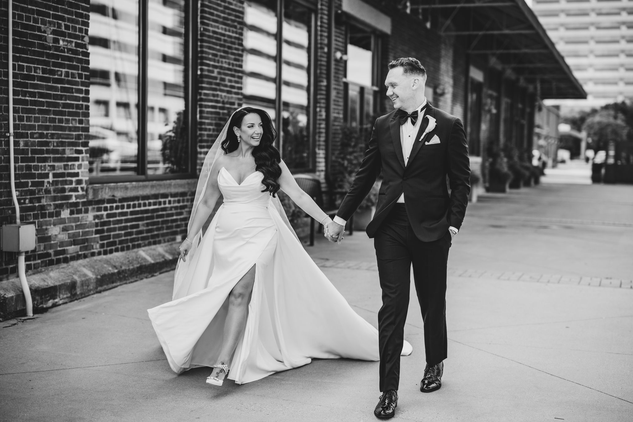 Wedding couple walks along brick building with urban architecture in background.