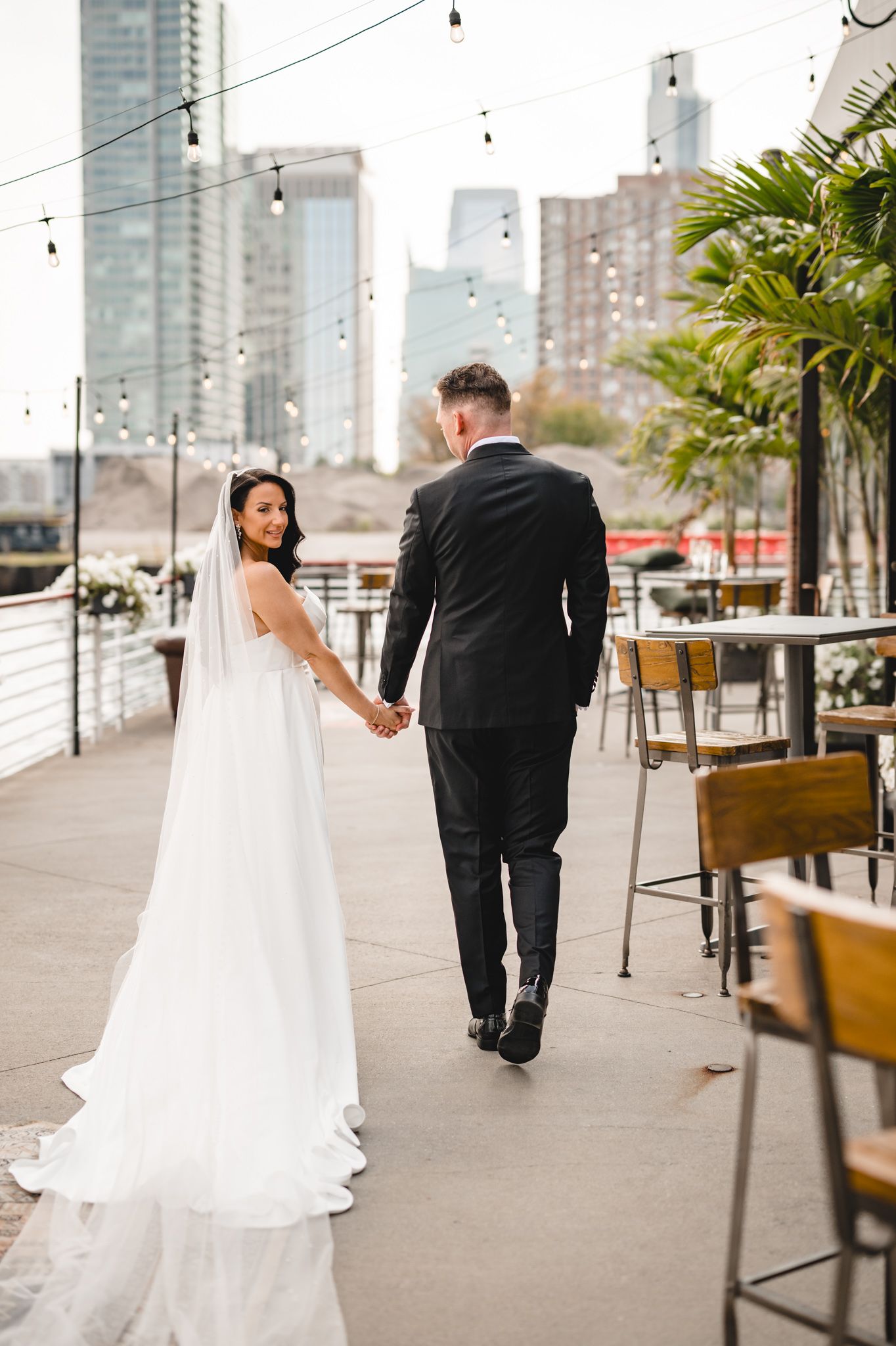 Bride and groom walk along waterfront promenade with modern cityscape in background.