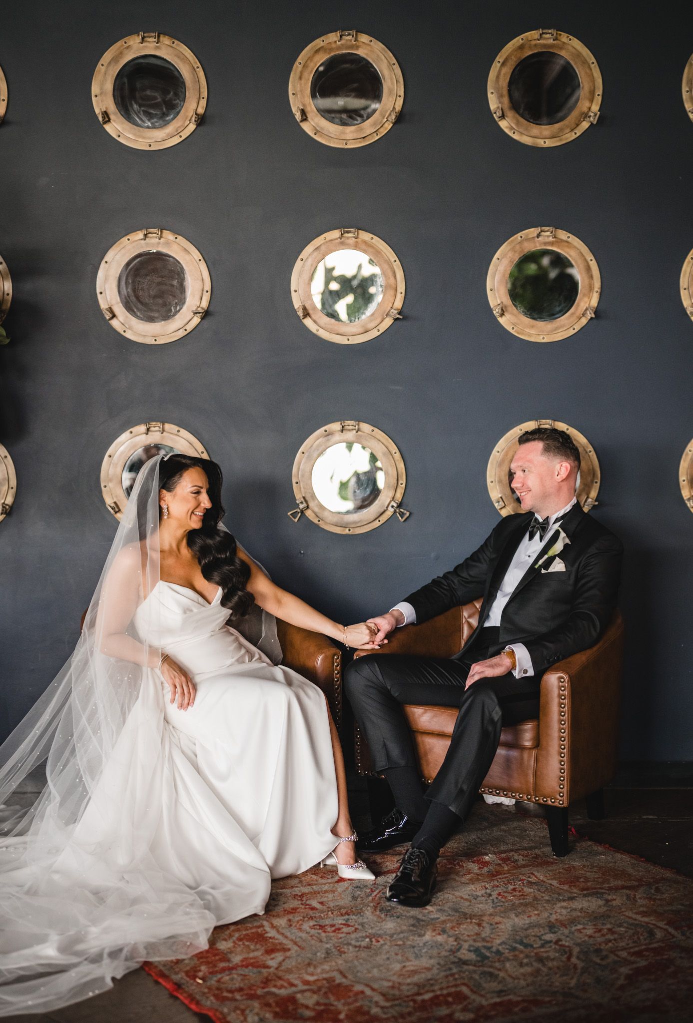 Stylish couple poses playfully on vintage furniture against wall with circular porthole decorations.