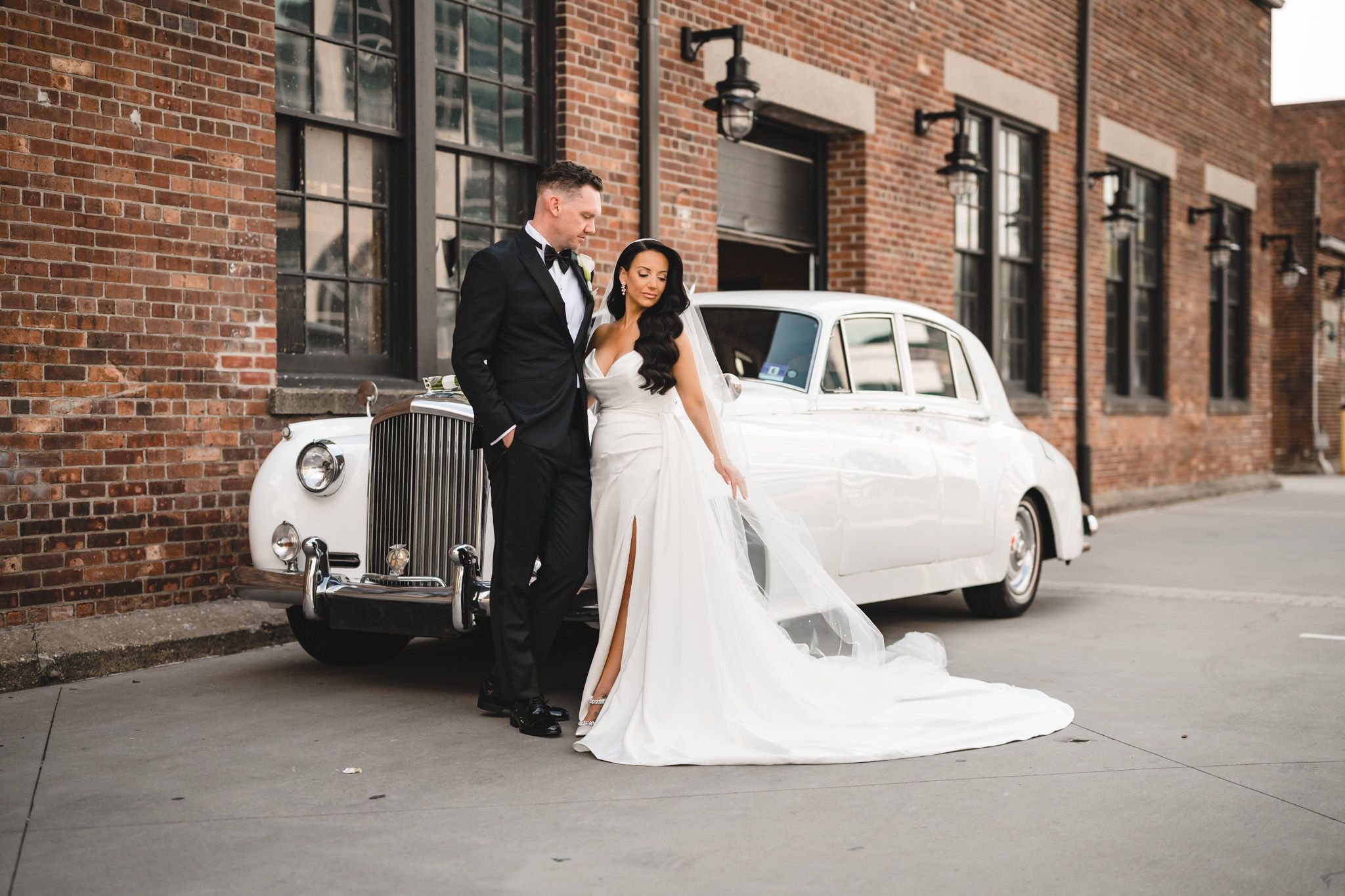 Wedding couple poses along brick wall with classic white vintage car in urban setting.