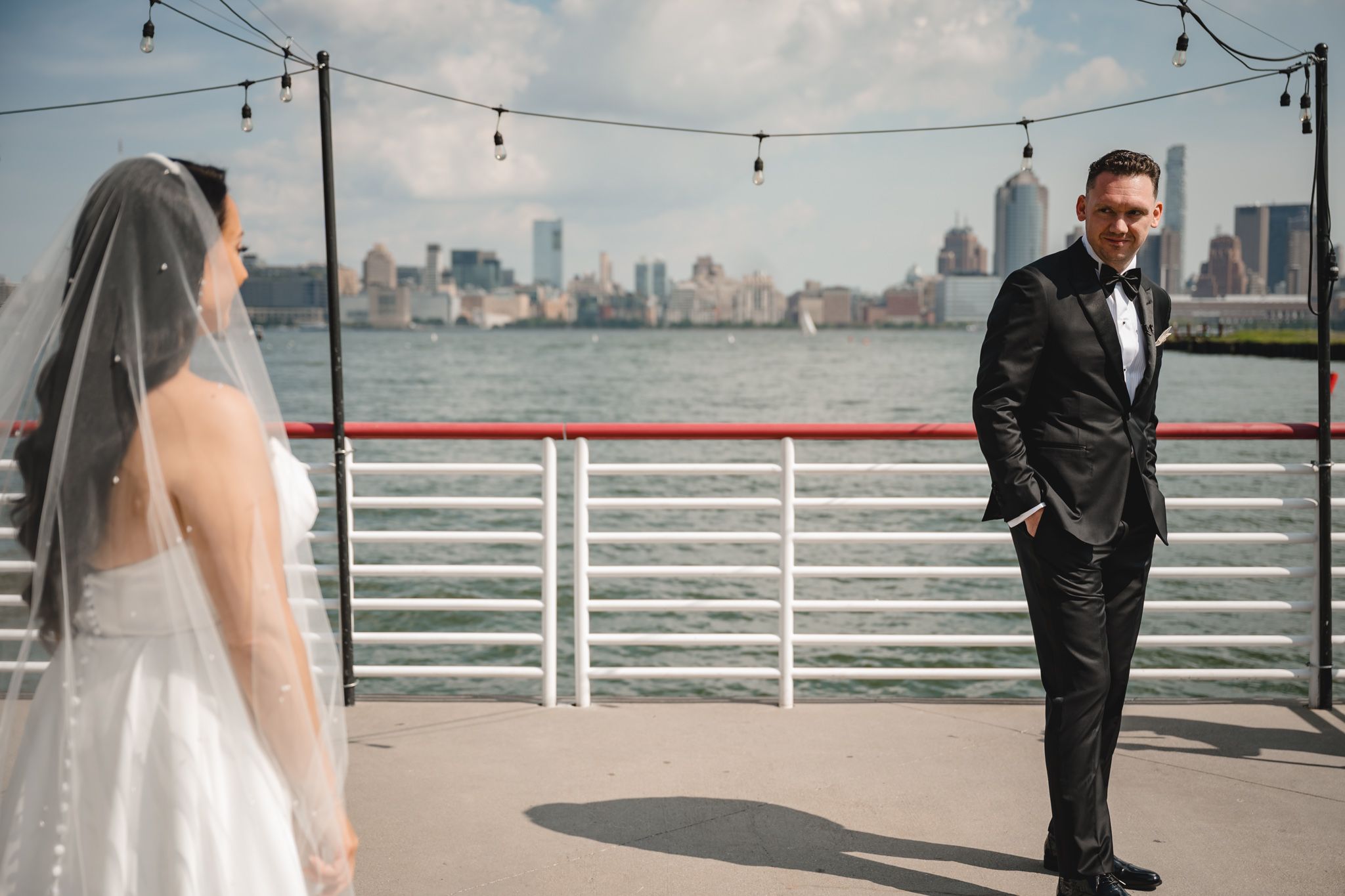 Wedding couple shares a romantic moment on a pier with string lights and city skyline.