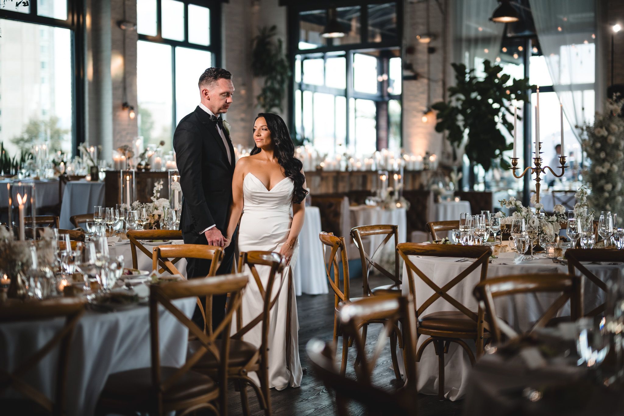 Elegant wedding reception venue with rustic wooden chairs and tables set for dinner under pendant lighting.