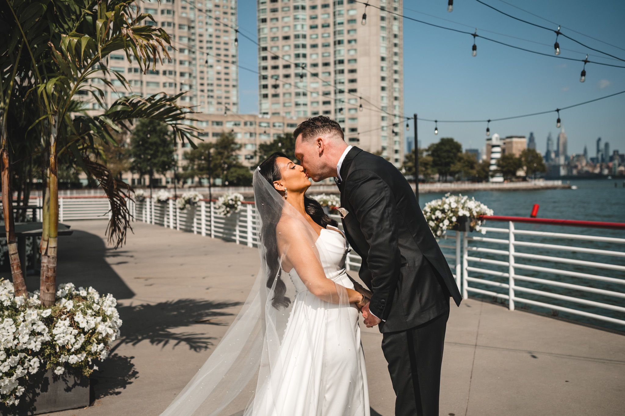 Bride and groom embrace on waterfront deck with Manhattan skyline visible.