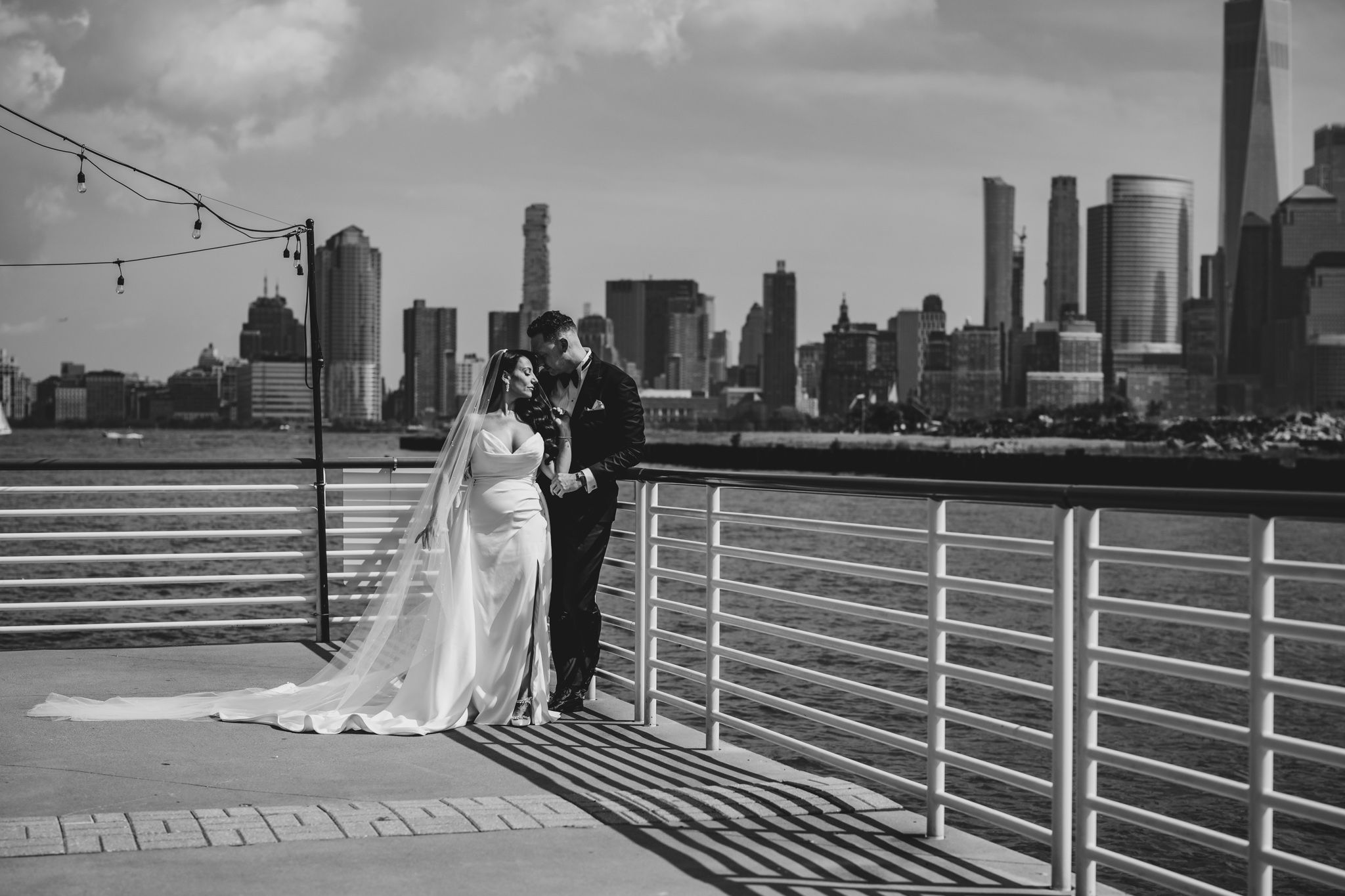 Black and white wedding photos on pier with city skyline backdrop.