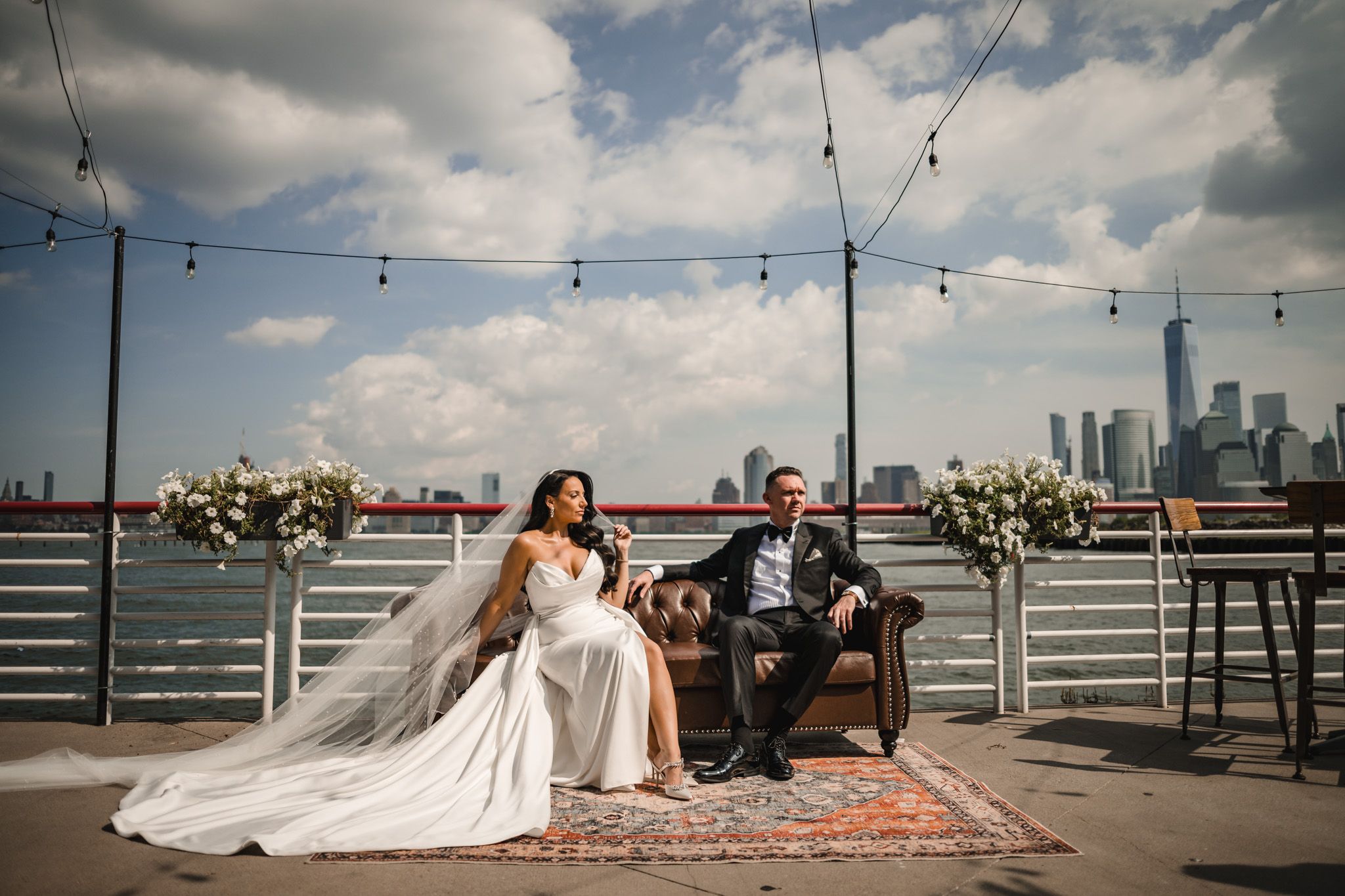 Wedding couple relaxes on vintage rug with string lights and cloudy sky overhead.