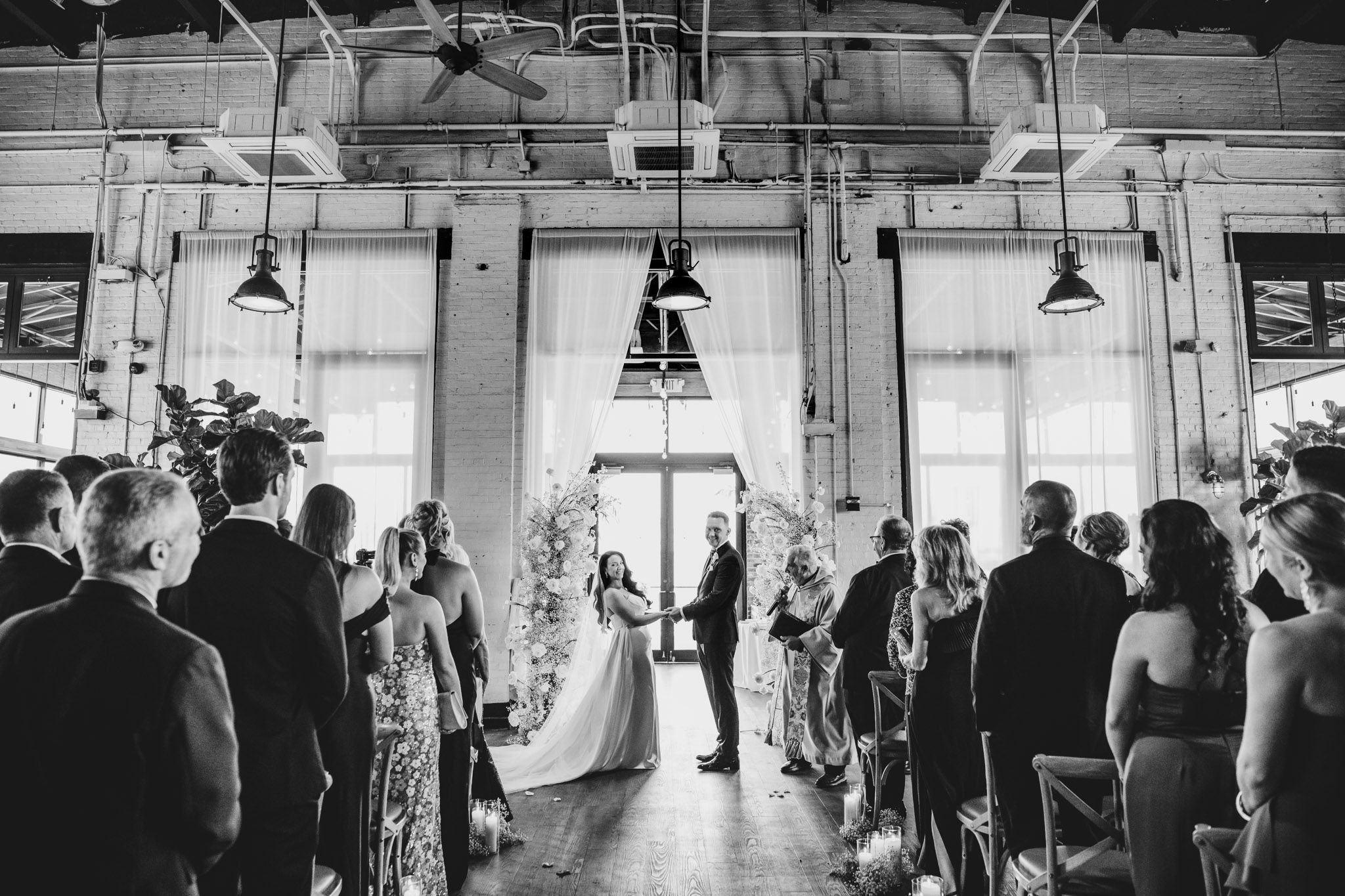 Black and white wedding ceremony photo showing guests watching couple exchange vows under industrial lighting.