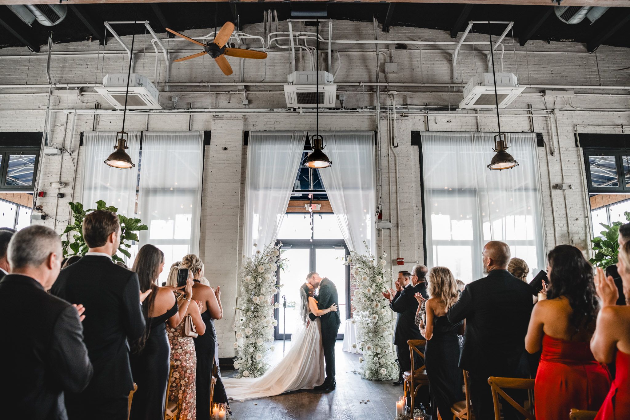 Wedding ceremony in a modern industrial space with white floral arch and pendant lighting.