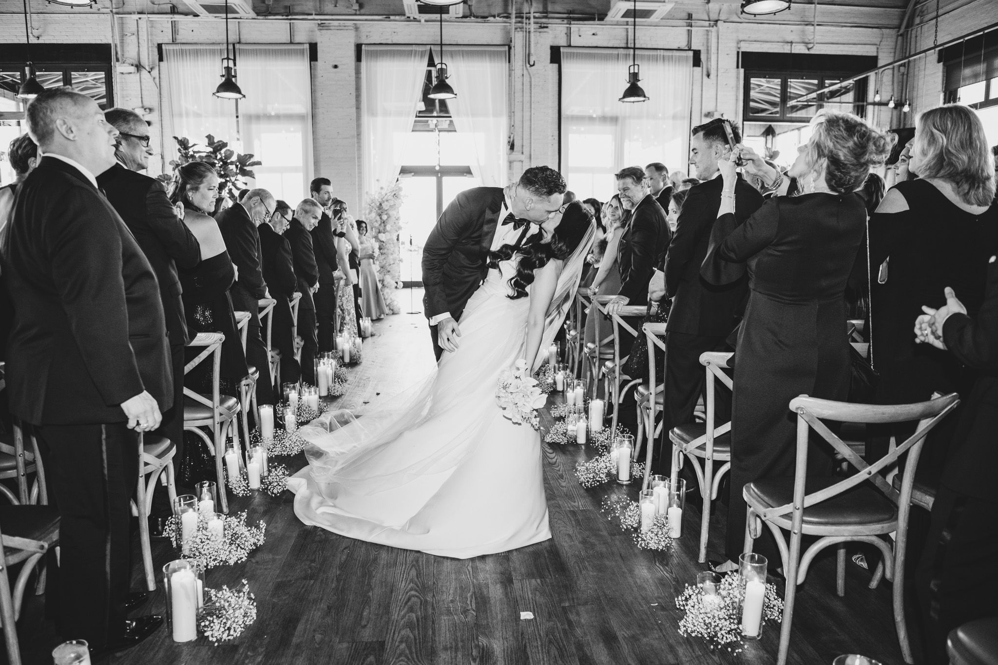 Black and white photo of wedding ceremony aisle lined with candles and cross-back chairs.