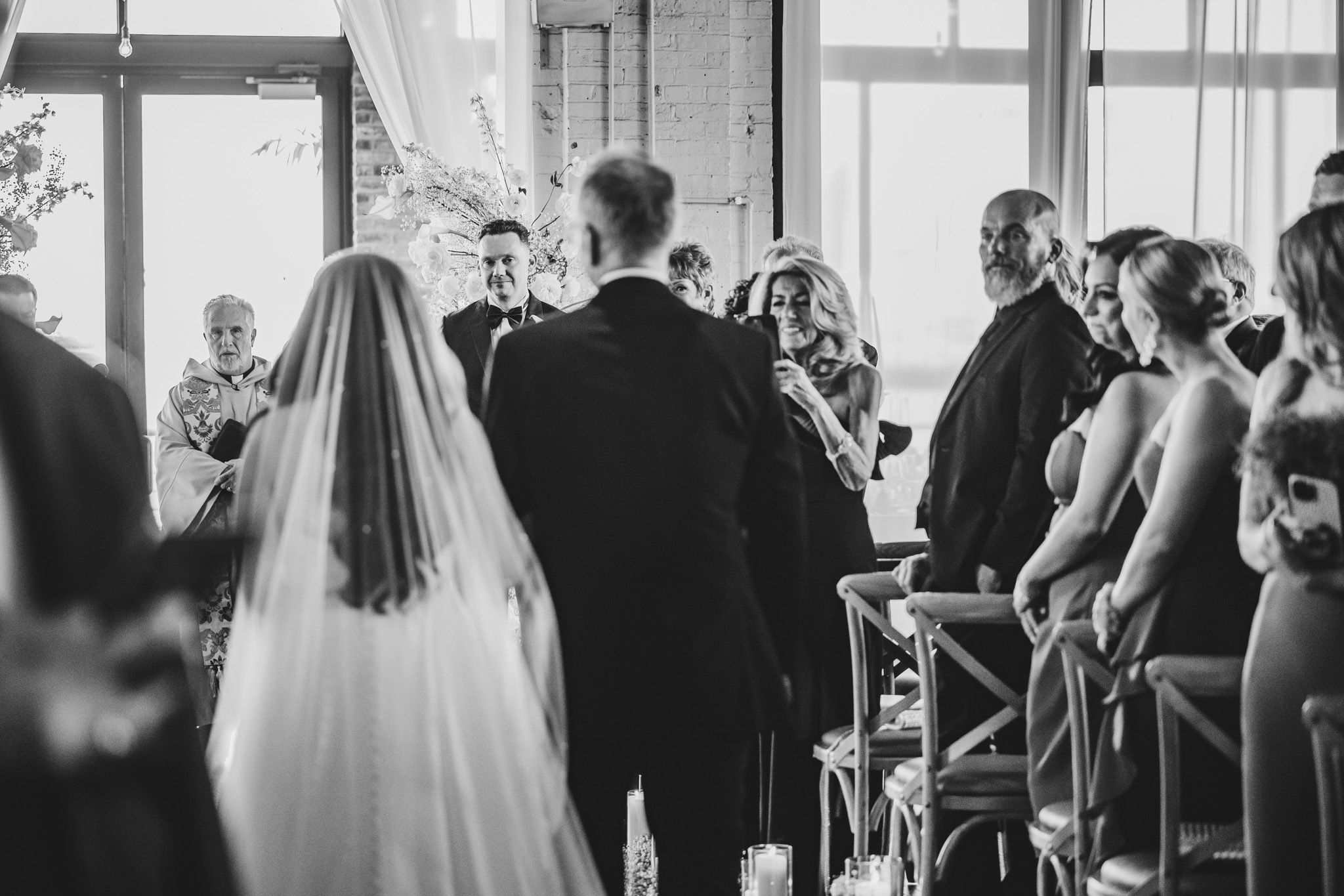 Wedding ceremony procession with guests standing in modern venue with large windows.