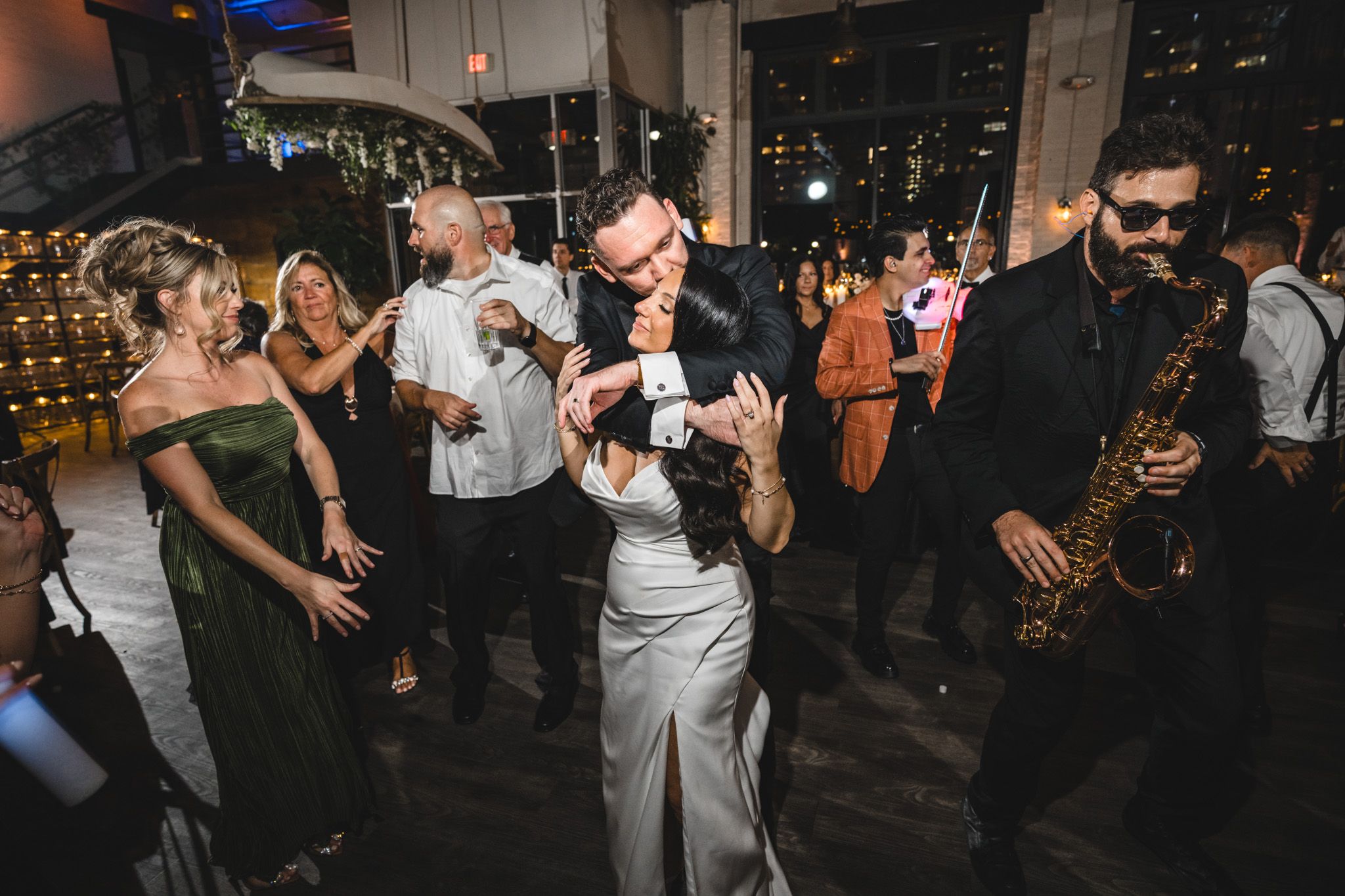Wedding guests dance and socialize during a lively reception with city lights in background.