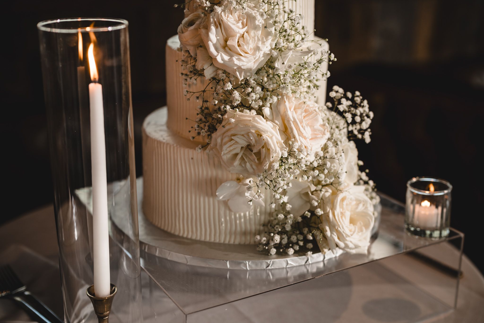 White wedding cake decorated with fresh roses and baby's breath alongside tall taper candles.