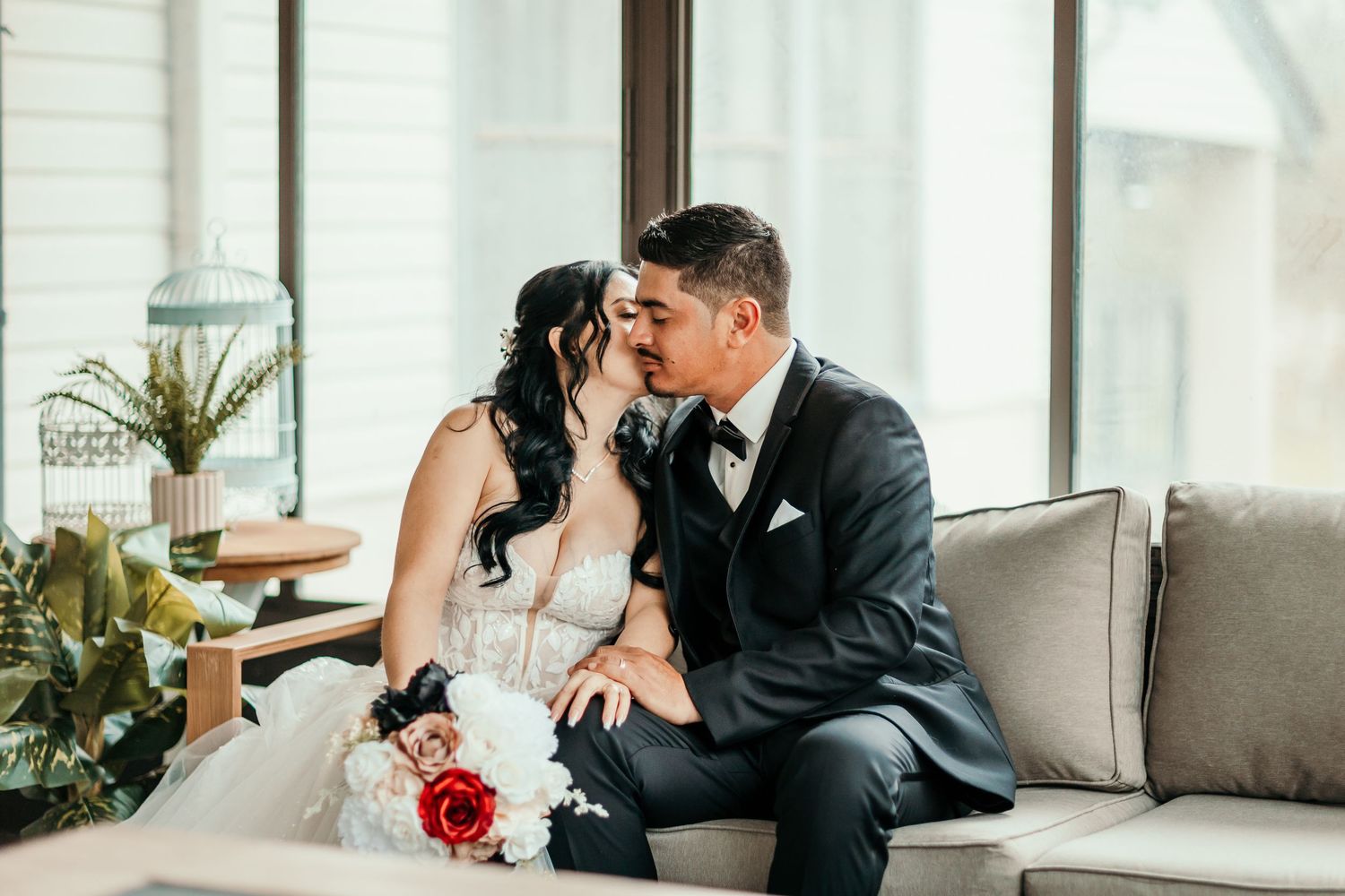 Bride and groom sharing a kiss while seated on a couch, bride holding a red and white bouquet.