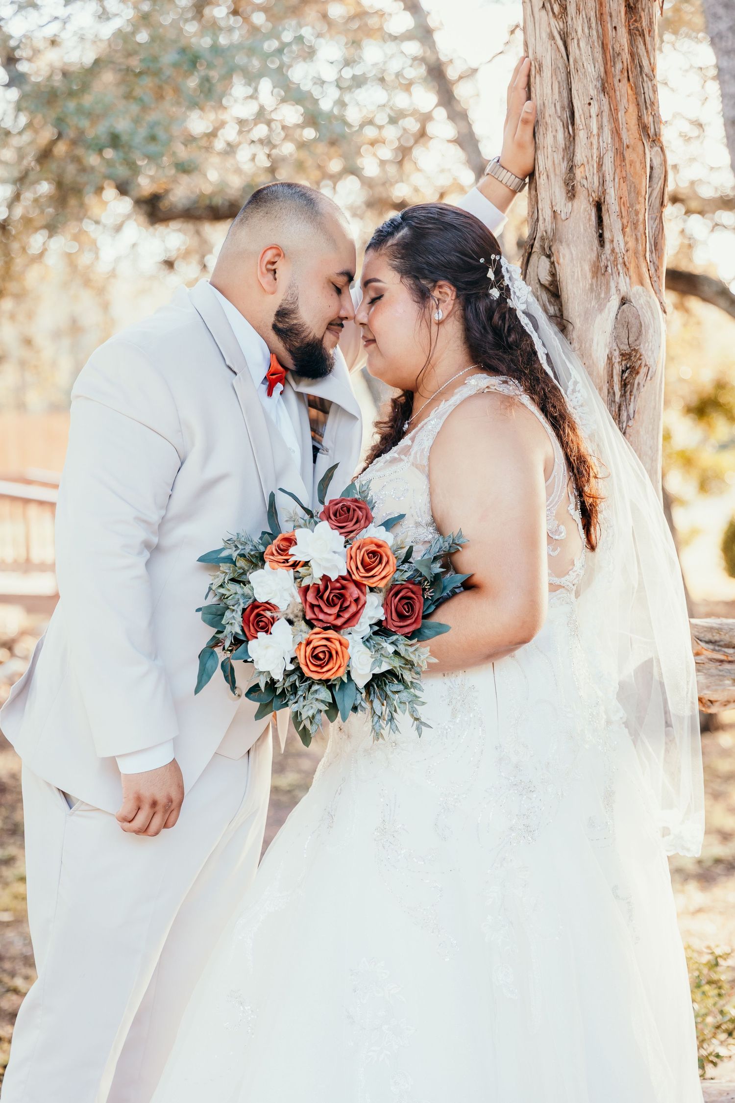 Bride and groom touching foreheads near a tree, bride holding a bouquet of red and white roses.