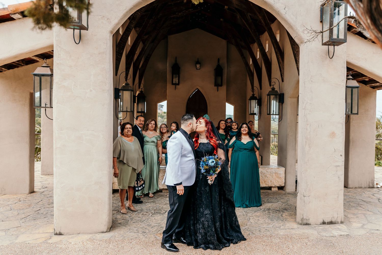 Bride in black lace gown kissing groom under stone arch with wedding party behind.