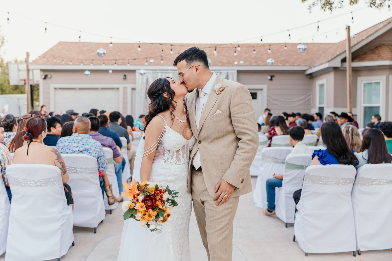 Bride and groom sharing a first kiss at an outdoor wedding ceremony with guests seated behind them.