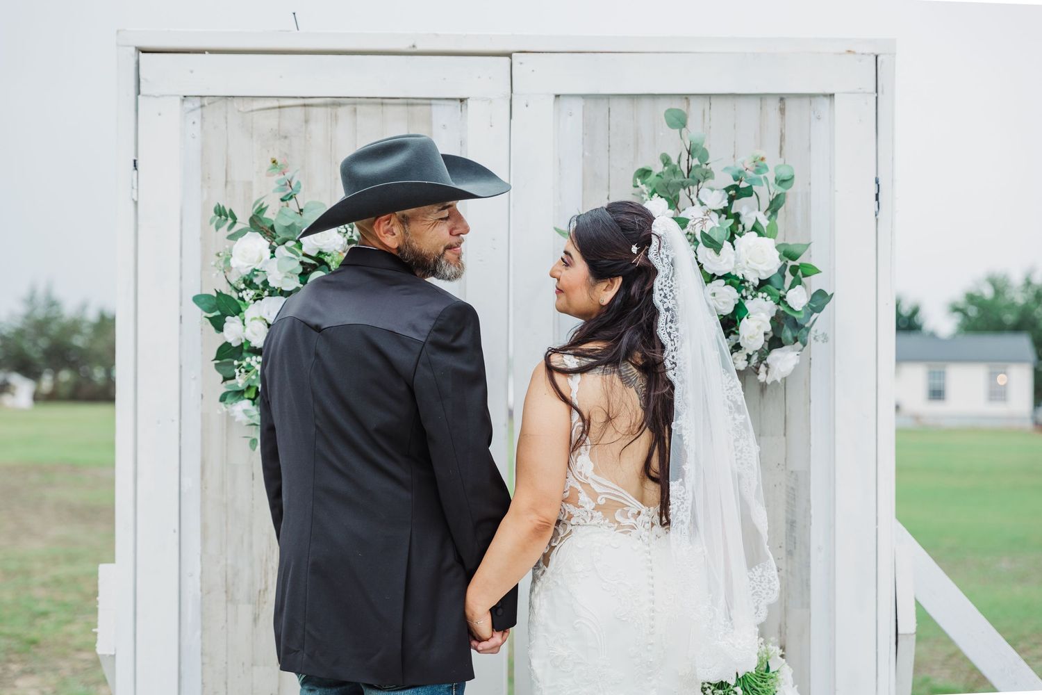 Bride and groom holding hands before a white door arch adorned with white flowers, groom in cowboy hat.