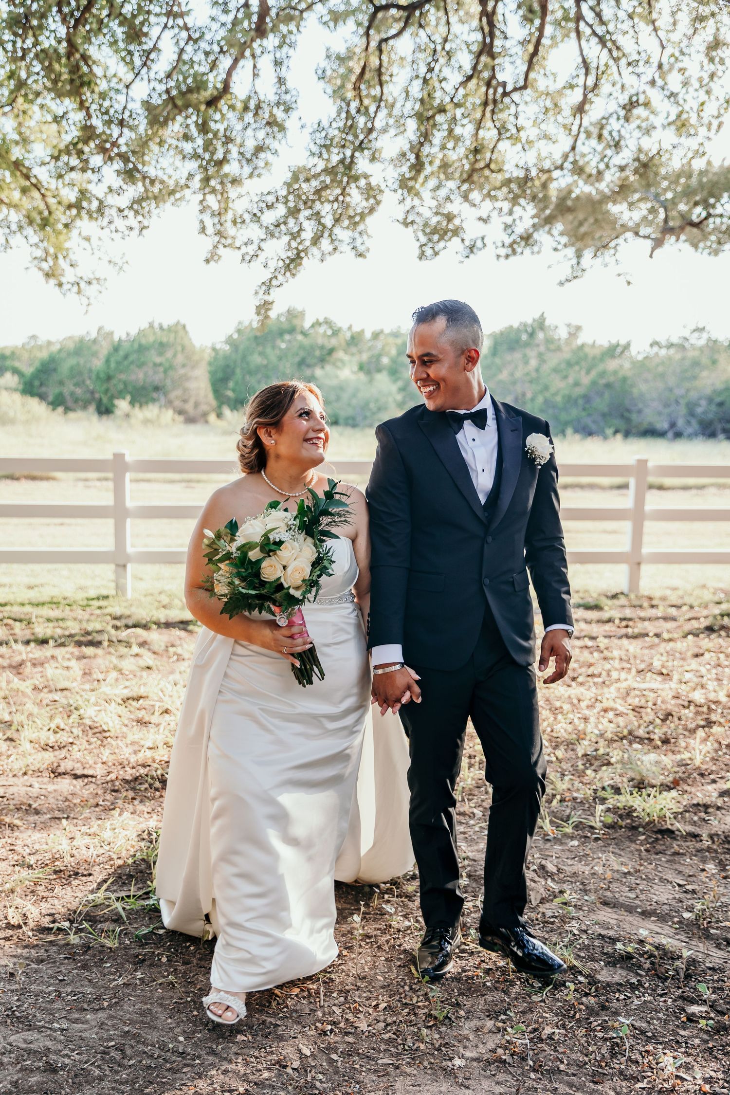 Newlywed couple walking hand in hand at rustic outdoor wedding venue with white fence.