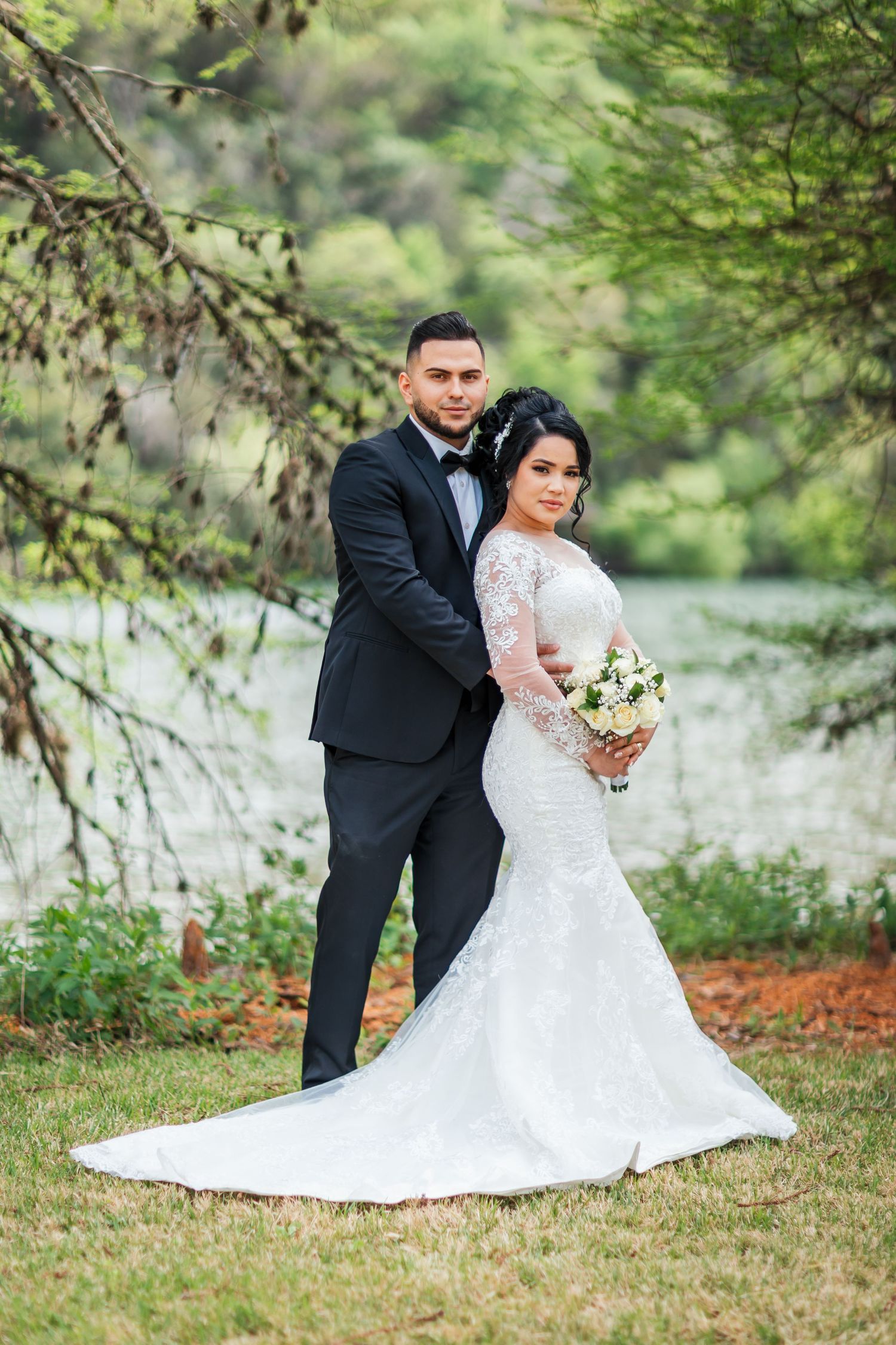 Bride in lace mermaid gown and groom in black tuxedo posing near a lakeside tree.