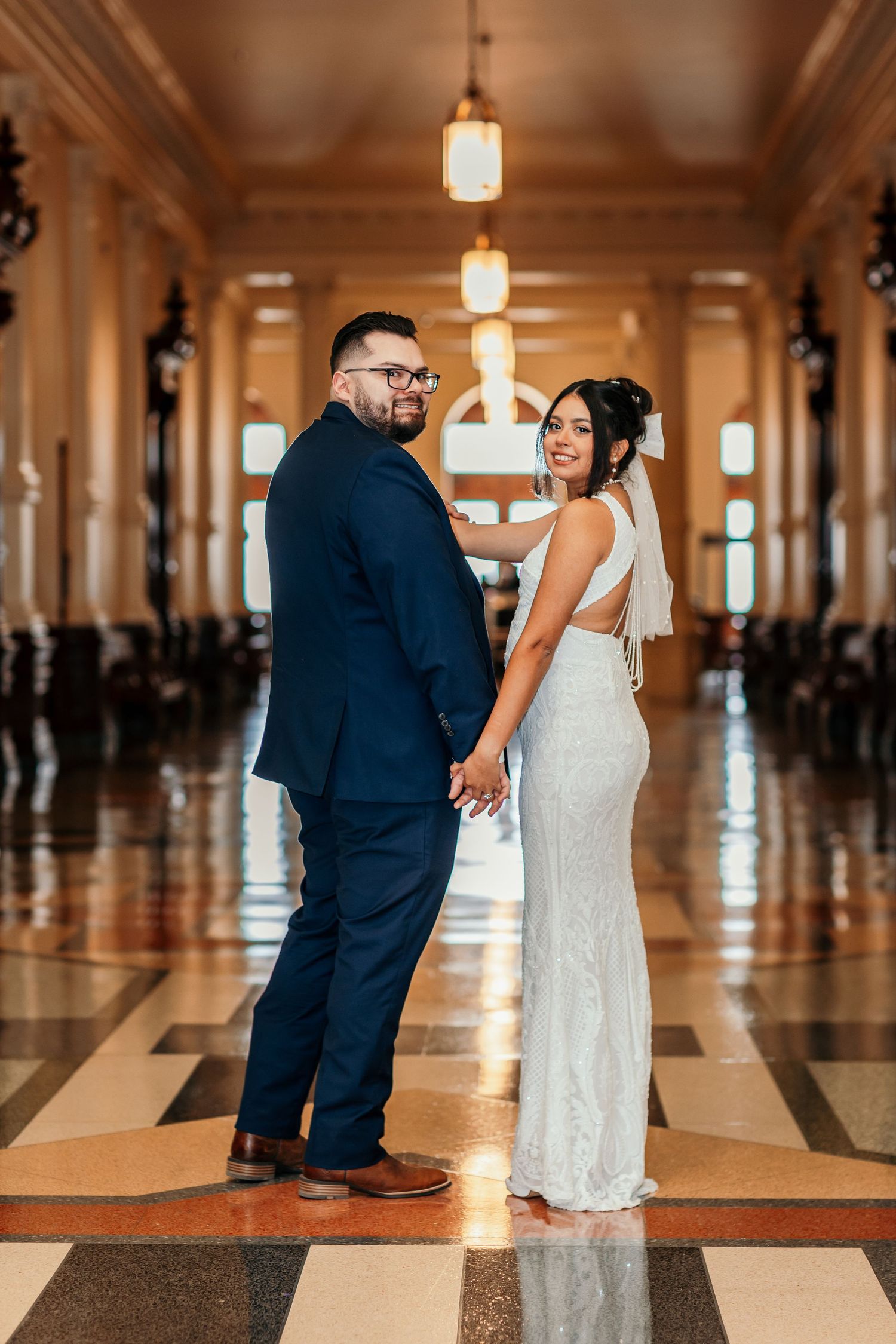 Bride and groom holding hands in an elegant grand hall with marble floors and ornate columns.