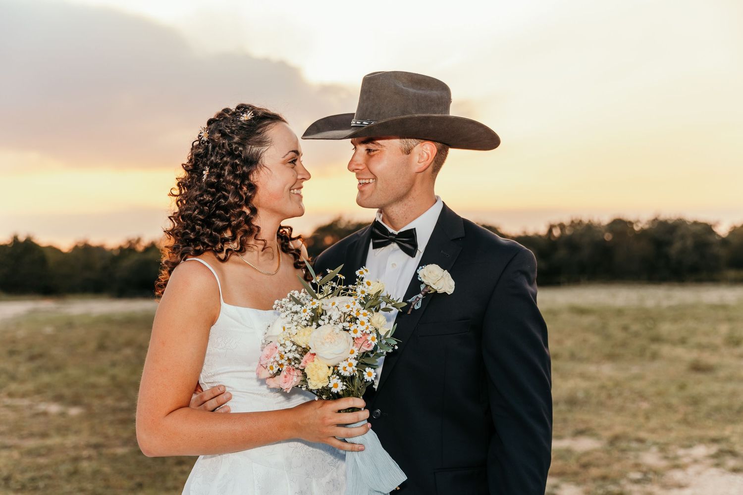 Bride and groom sharing a romantic moment at sunset in an open field, groom wearing a cowboy hat.