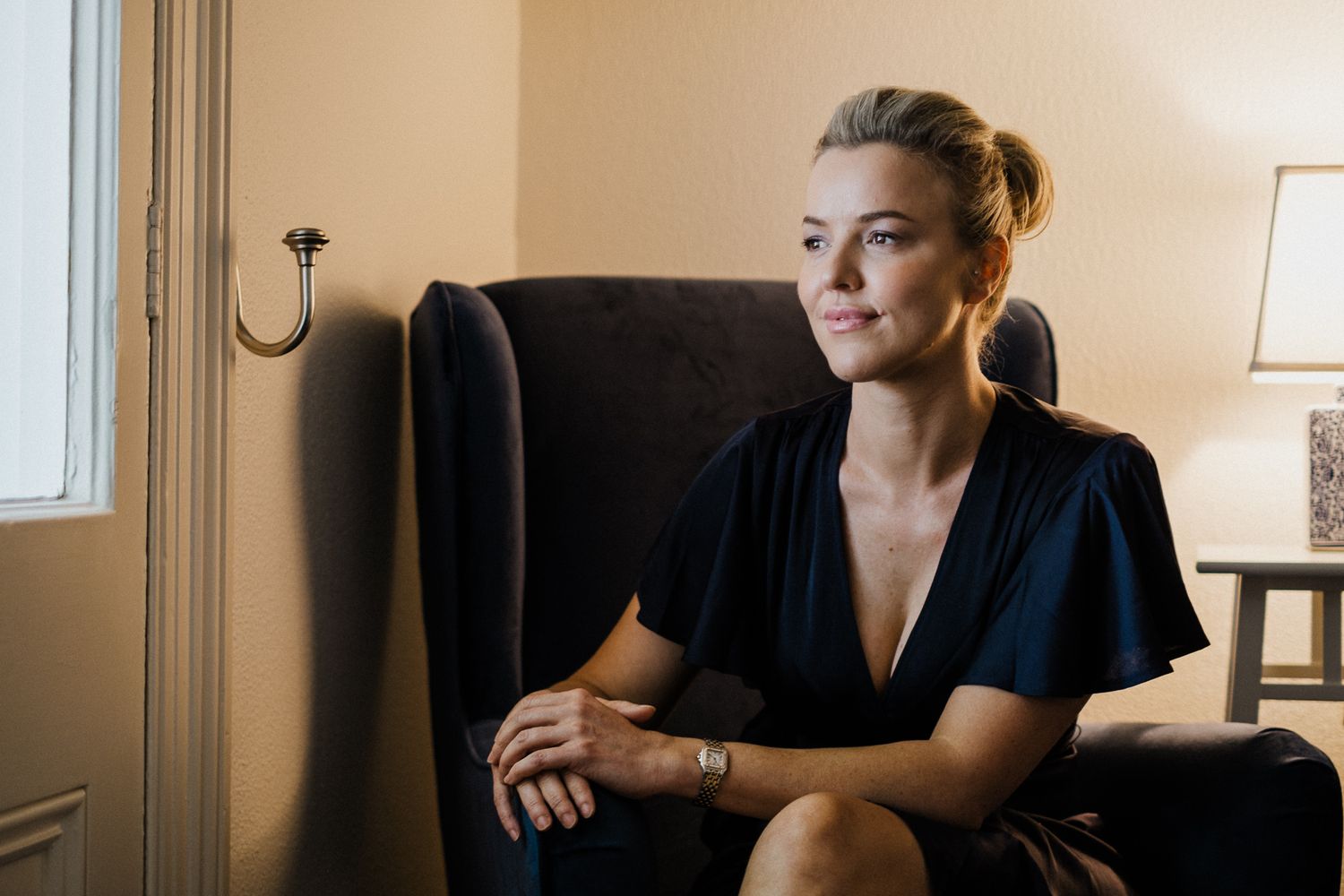 A person in a black top sits on a couch in a dimly lit room, looking thoughtfully to the side.